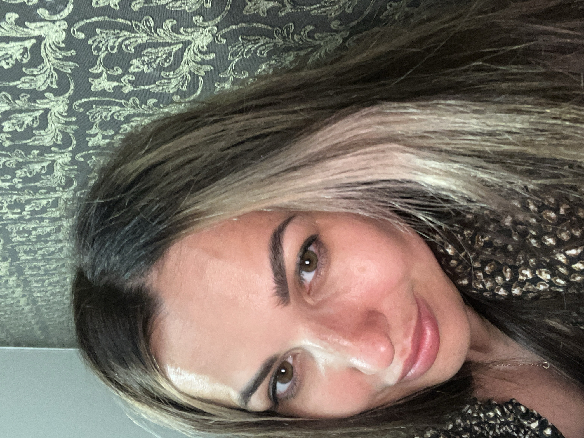 A woman with brown hair and brown eyes is shown in a close-up shot. She is wearing a patterned top and a silver necklace. The background is a patterned wallpaper.