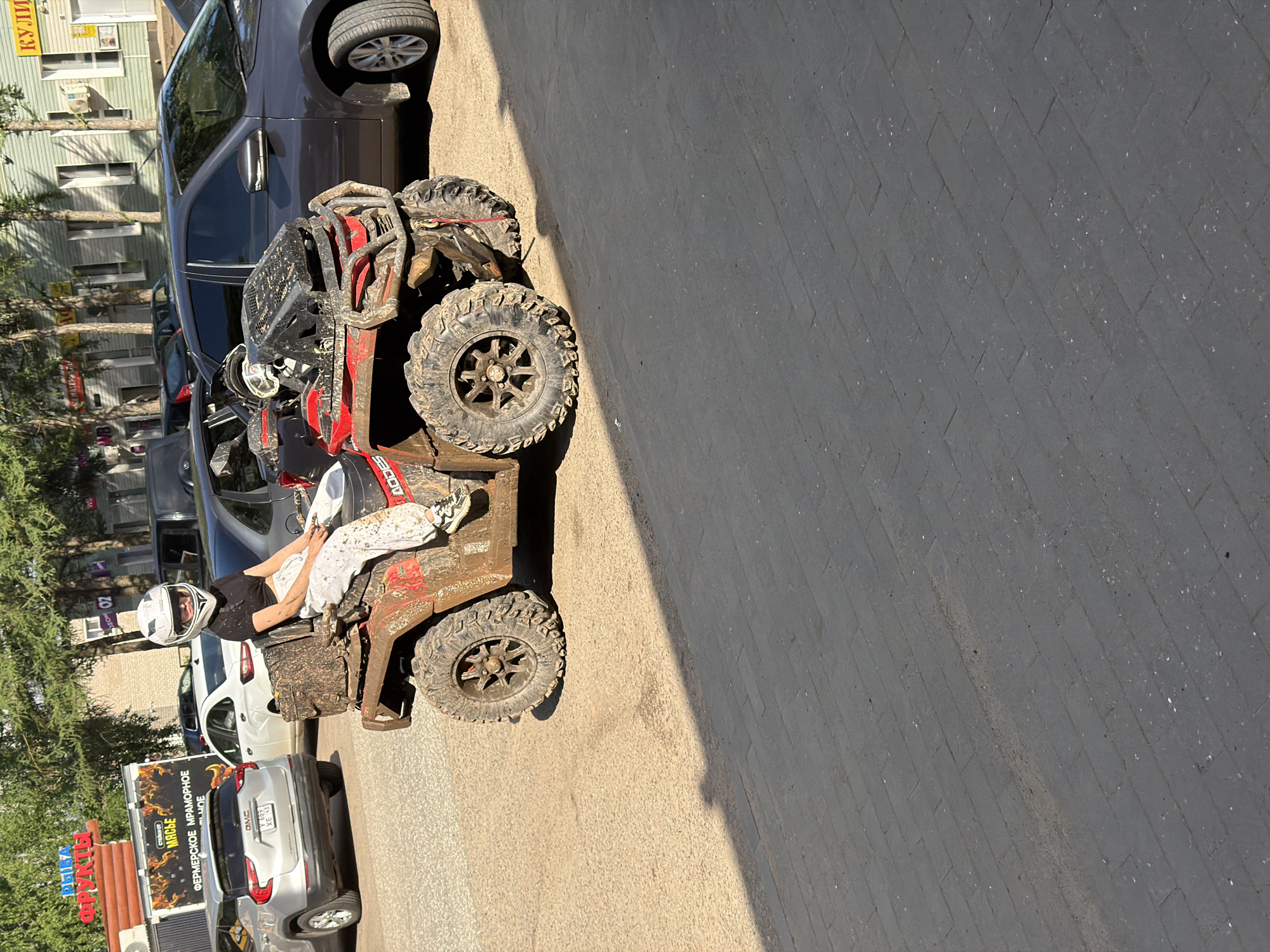 A young woman wearing a white helmet and white pants is sitting on a mud-covered red ATV. The ATV is parked on a dusty surface. In the background, there are cars, buildings, and trees. One of the buildings has a sign that reads "ФРУКТЫ" (Fruits) and another has "КУЛИ" (Kulí). A silver GMC SUV is parked to the left of the ATV, and a dark-colored sedan is parked behind it. The scene appears to be in an outdoor urban environment.
