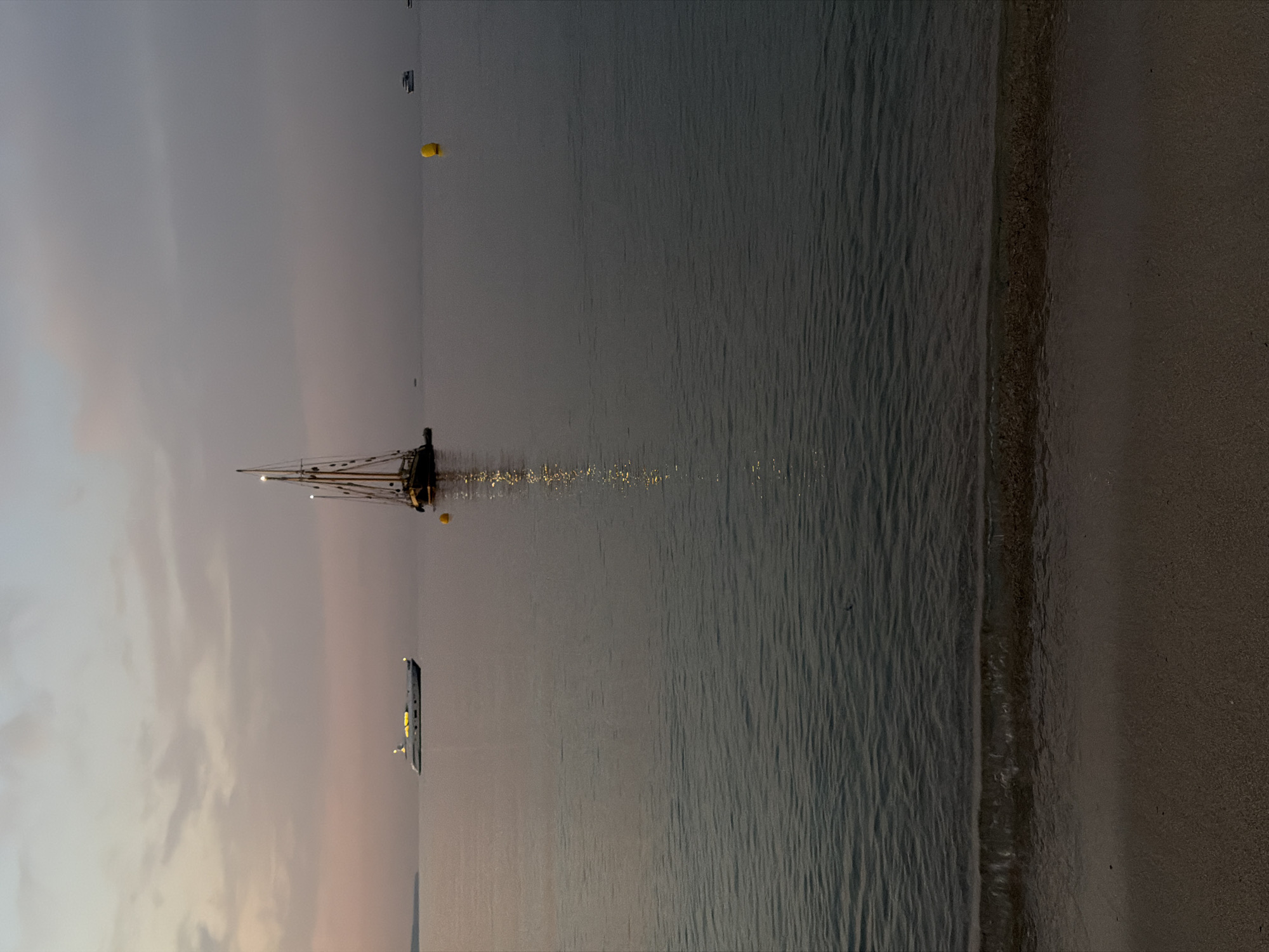 The image depicts a serene seascape at dusk, likely near Calvià, Spain, given the Mediterranean aesthetic. The foreground features a sandy beach with gentle waves lapping at the shore. The sea itself is calm and reflects the dusky sky, which is painted with muted tones of pink and gray. In the mid-ground, several vessels are anchored. A modern white yacht with illuminated windows is visible on the left, suggesting a luxurious setting. Further to the right, a traditional sailing ship with multiple masts stands out, its rigging silhouetted against the fading light. A bright yellow buoy floats in the water between the two larger vessels. The horizon is indistinct, merging the sea and sky into a soft, atmospheric haze. The overall impression is one of tranquility and the peaceful end of a day by the coast.
