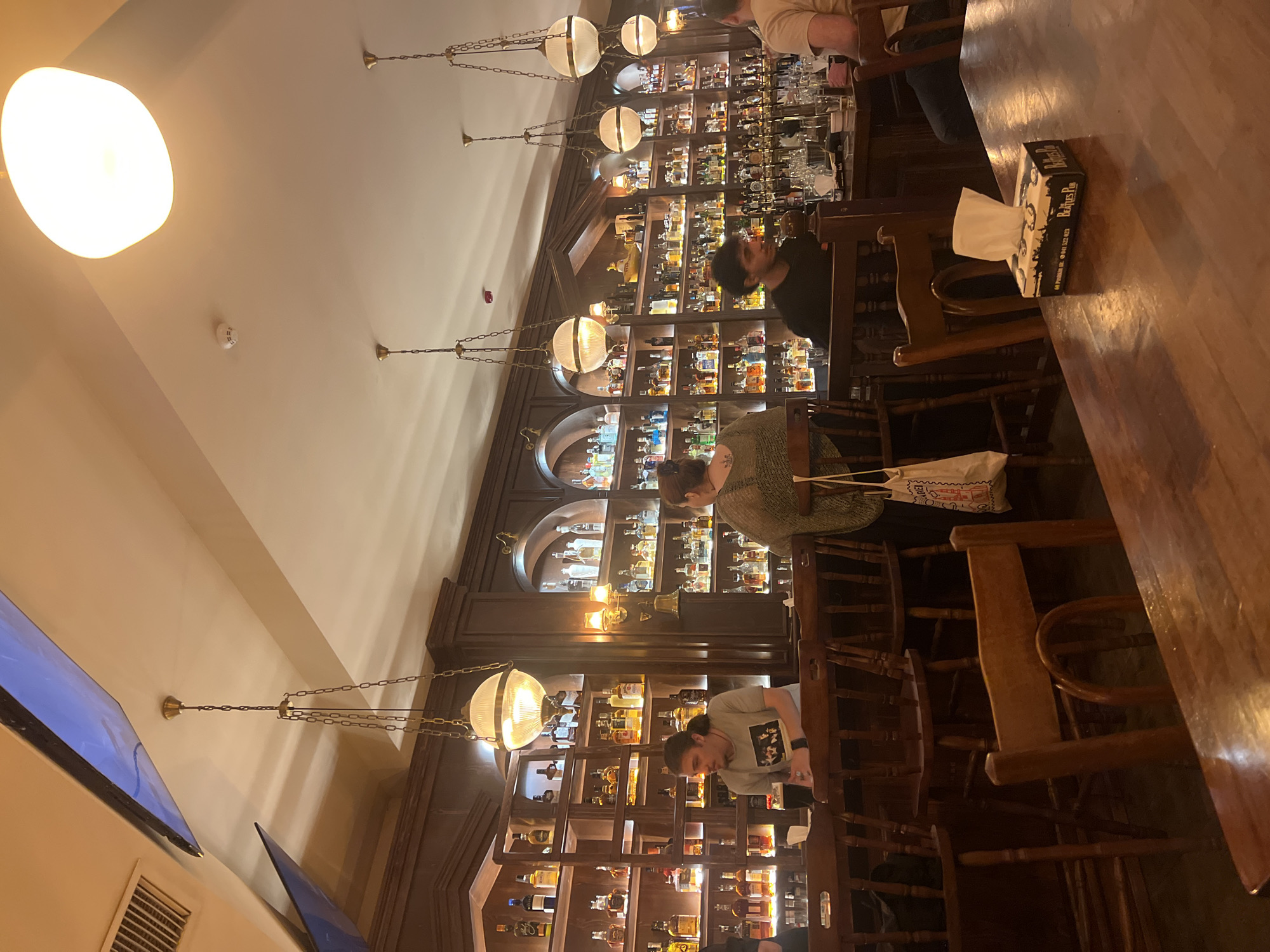 The image shows the interior of the Beatles Pub in Yerevan, Armenia. A bartender, a man with long hair tied back, stands behind a wooden bar, facing to the left. He is wearing a grey t-shirt with a graphic on it. To his right, a woman with dark hair is seated at the bar, facing away from the camera, wearing a green knit sweater. Further down the bar, another person, a man with dark hair, is partially visible, wearing a black shirt. He appears to be interacting with a customer on the far right of the image, whose back is to the camera. The bar is lined with shelves filled with numerous bottles of alcohol. Several pendant lights with spherical glass shades hang from the ceiling, illuminating the space. Wooden chairs are positioned around the bar and at a table in the foreground. A box of tissues, branded "Beatles Pub," sits on the wooden table in the foreground. The overall ambiance is warm and dimly lit, suggesting an evening setting.