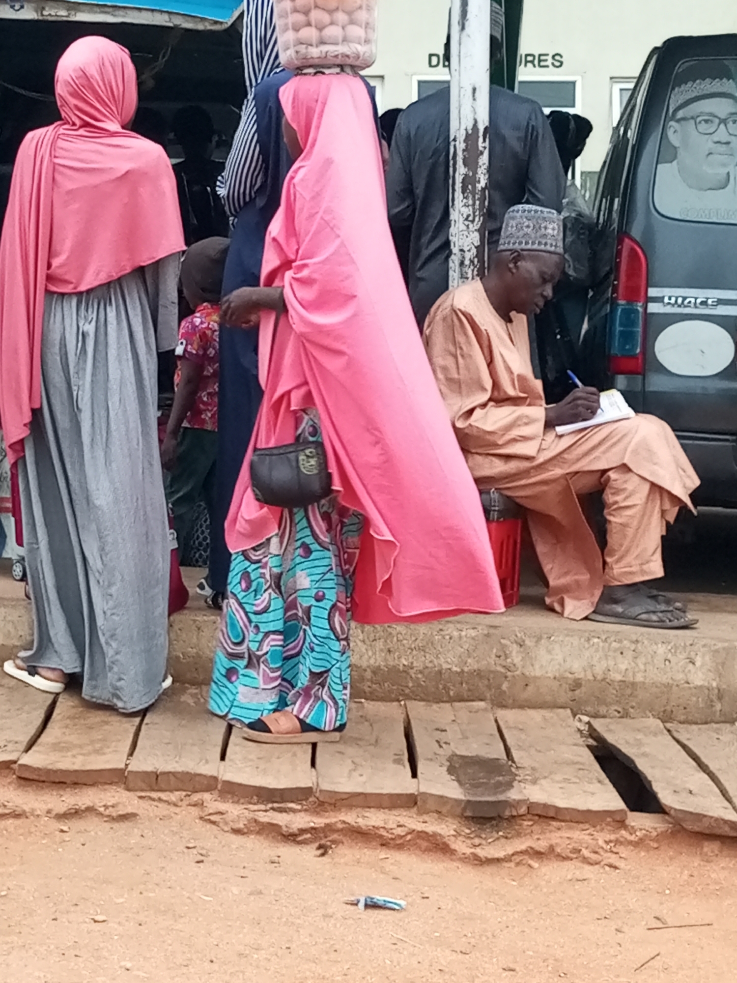 The image captures a scene in Gombe, Nigeria, likely outdoors, with a group of people gathered around a temporary stall or market area. The time of day appears to be daytime, possibly late morning or early afternoon, given the lighting. The weather seems to be warm, as evidenced by the light clothing worn by some individuals and the general setting.
The foreground is dominated by a wooden platform or walkway over loose soil. To the left, a person in a long grey skirt and pink headscarf is standing, their feet clad in white sandals. Beside them, another person is mostly obscured, but appears to be wearing a pink headscarf and a vibrant blue and pink patterned skirt. This individual also has a small dark handbag slung over their shoulder. Further back, a child wearing a brightly colored shirt is visible.
To the right, an older man is seated on a concrete step, wearing an orange traditional outfit and a patterned cap. He is actively writing in a small notebook with a blue pen. Behind him, a dark colored van, possibly a Toyota Hiace, is parked. The van has a large black and white sticker on its rear window featuring a portrait of a man in glasses. Some text is visible on the van, including "HIACE" and what appears to be a partial inscription below the portrait.
In the background, more people are standing, and a building with signage can be seen. The partial text on the building reads "DE" and "RES", suggesting a business or office. The overall impression is one of everyday life and commerce in a Nigerian setting. The man writing appears focused on his task, while the surrounding individuals are engaged in activities related to the market or waiting. There are no strong indicators of emotion, but the scene suggests a relatively calm and organized environment.