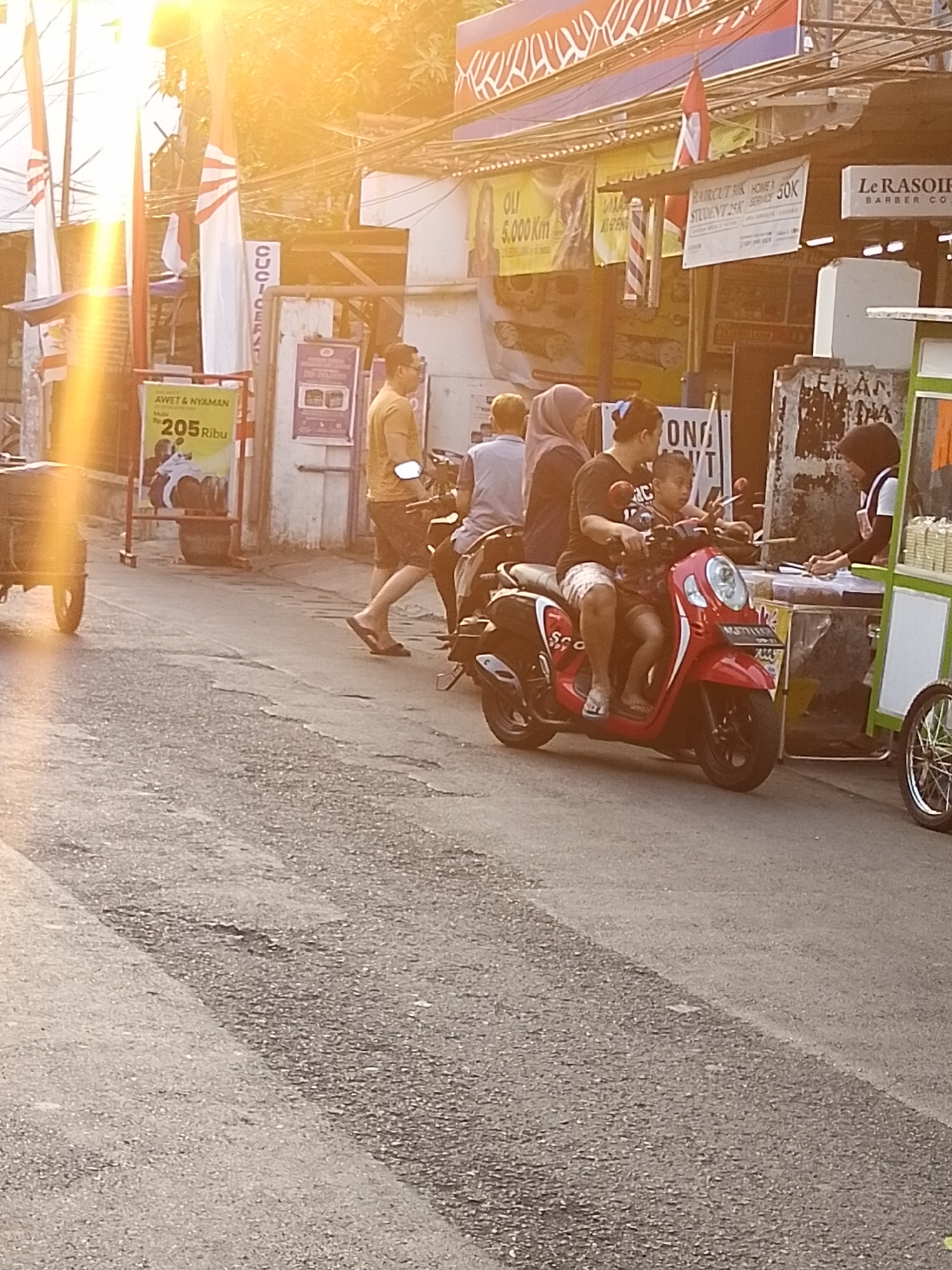 The image depicts a street scene in Surabaya, Indonesia, likely during the late afternoon or early evening, as indicated by the warm, golden sunlight. The street is paved with asphalt and shows signs of wear. Several people are present, engaged in various activities.
In the foreground, a red scooter is parked, carrying a woman and a young child. The woman appears to be driving, and the child is seated in front of her. Both are looking forward. To the right of the scooter, a street vendor operates a cart, which is green and white, selling what appear to be food items in containers. A woman, possibly the vendor, is interacting with customers.
Behind the scooter, several other individuals are visible. A man in a yellow shirt and dark shorts is walking away from the camera, wearing flip-flops. Another person, in a light blue shirt, is partially visible behind him. Further in the background, there is a suggestion of buildings and shops lining the street, adorned with various banners and signs, some of which appear to be advertisements. One sign displays "Le Rasoir BARBER CO." suggesting a barber shop. Another sign promotes "AWET & NYAMAN" (Durable & Comfortable) with a price of "205 Ribu" (205 thousand). Indonesian flags are also visible, mounted on poles. The overall atmosphere suggests a busy urban environment with typical street life.