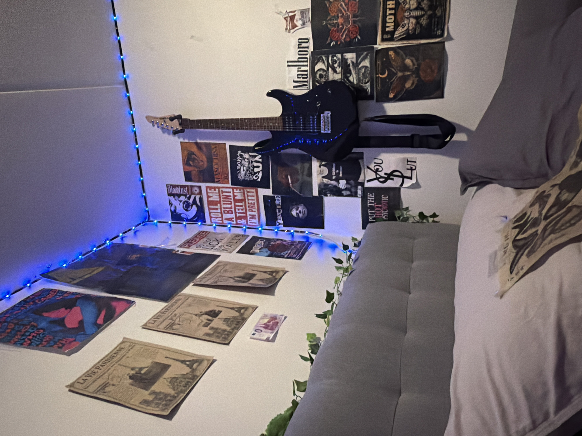 The image depicts a bedroom with a minimalist aesthetic, characterized by white walls adorned with various posters and a black electric guitar mounted on the wall. A string of blue LED lights runs along the ceiling corner, casting a blue hue on the scene. To the left of the image, a gray upholstered headboard and a pillow suggest a bed. A monetary note is affixed to the wall below several vintage-style posters featuring Parisian landmarks. The room appears to be a personal space, likely a teenager's bedroom, given the décor. No specific location in Oslo, Norway is identifiable from the provided image.