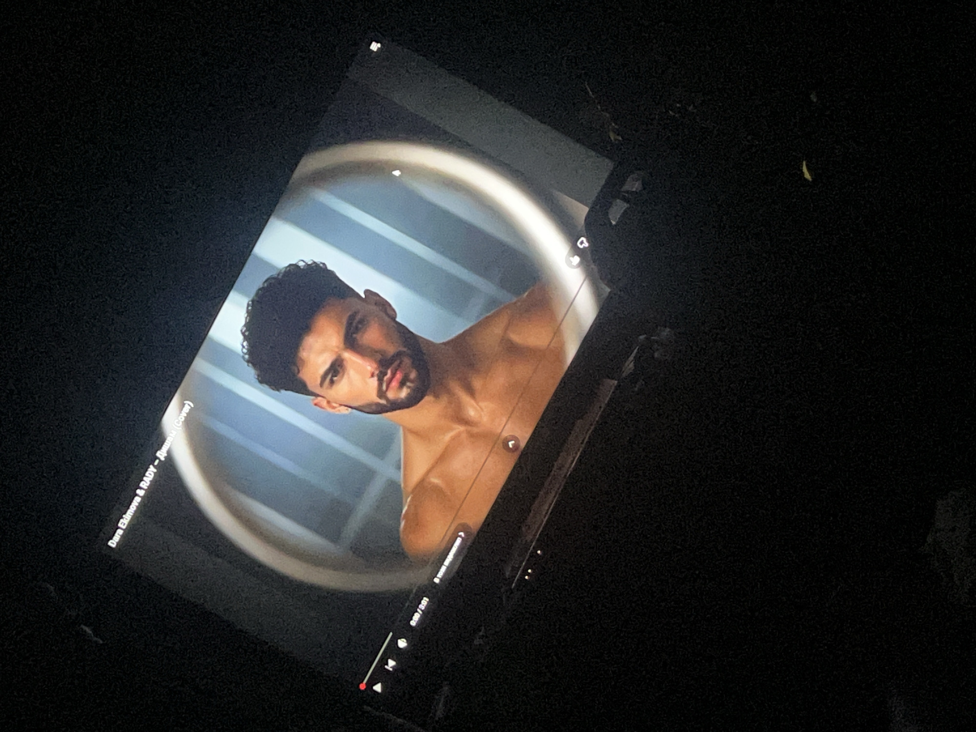 The image shows a screen displaying a music video by Dara Ekimova & RADY. The title of the song is "Дишам (Cover)". The video features a shirtless man with dark hair and a beard. The screen is tilted and surrounded by darkness. The video is playing on a device, possibly a tablet, with the video controls visible at the bottom of the screen. The video is 0:00 into the 3:01 length. The location, Cherven Bryag, Bulgaria, is not discernible from the image.
