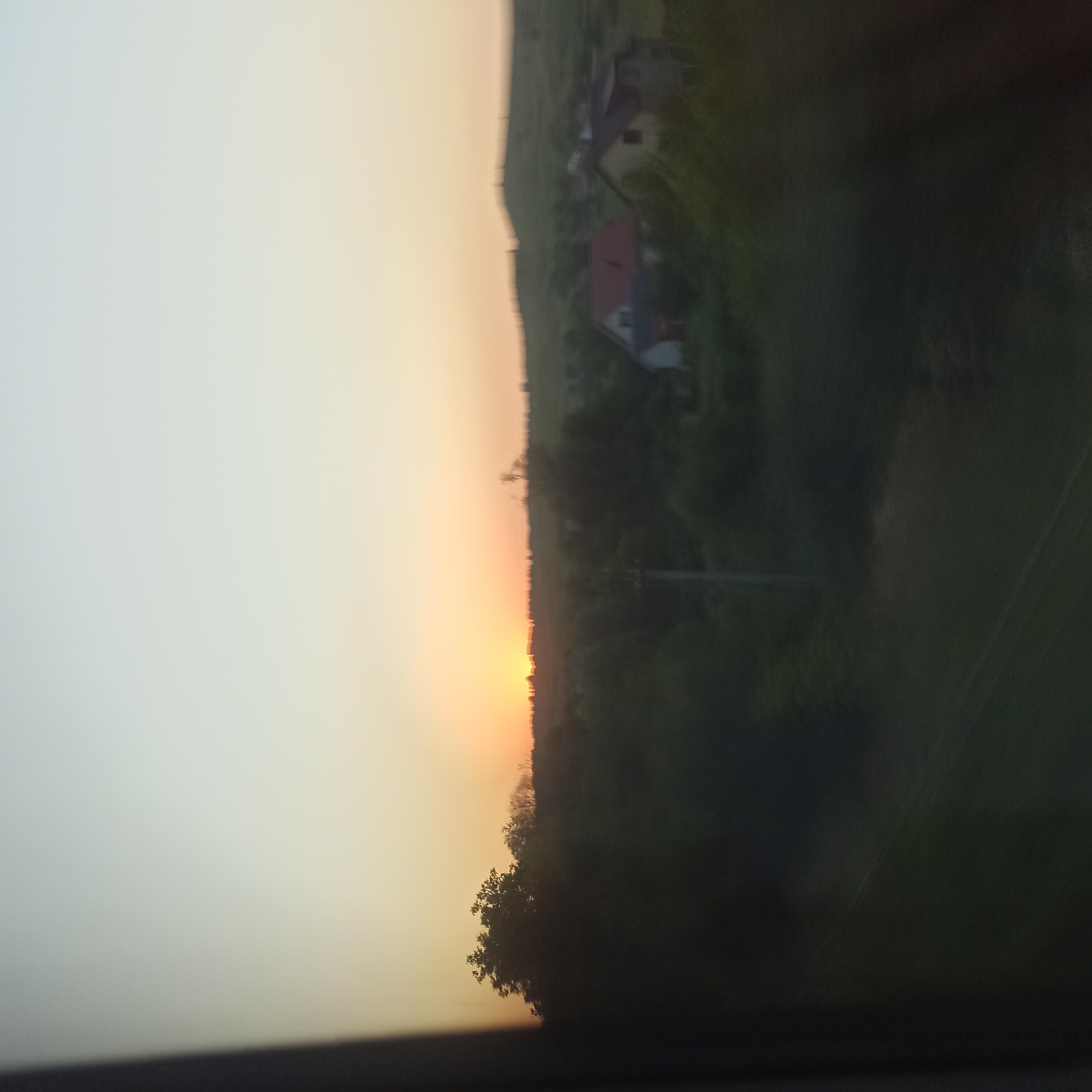The image captures a sunset over a rural landscape with the sun low on the horizon, casting a warm glow on the sky. In the foreground, a dark, blurry silhouette of trees and vegetation dominates the lower portion of the frame. In the middle ground, a few buildings with red and yellow roofs are visible, partially obscured by the foliage. The background features rolling hills and a distant treeline against the illuminated sky. The overall scene suggests a peaceful, natural environment. The image is taken from a slightly elevated, moving perspective, as indicated by the blur. The location is specified as Zavoloka, Ukraine.