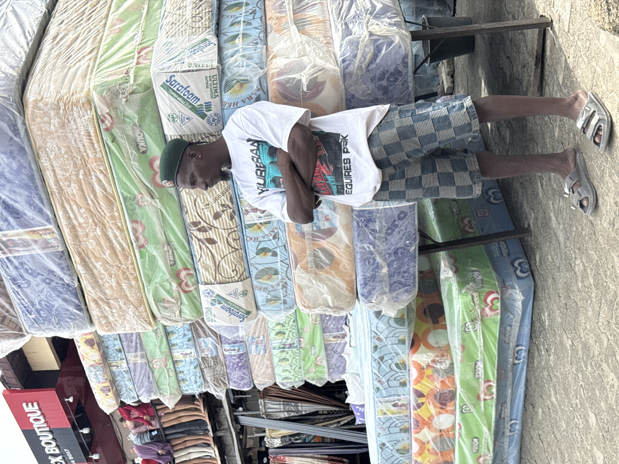 The image shows a street scene in Lagos, Nigeria with a man standing on a rack of mattresses. The mattresses are wrapped in plastic and stacked vertically. The mattresses feature brands such as "Sarafoam," "Ultima," and "Mouka" and show various decorative patterns. The man is wearing a white T-shirt with "Yureban Requires Pisk" printed on it, checkered shorts, a green beret, and sandals. His arms are crossed. In the lower left corner of the image is a red sign reading "EX BOUTIQUE". Behind the mattresses, racks of other items, perhaps clothing or textiles, are visible. The background shows a paved surface, likely a street or sidewalk.