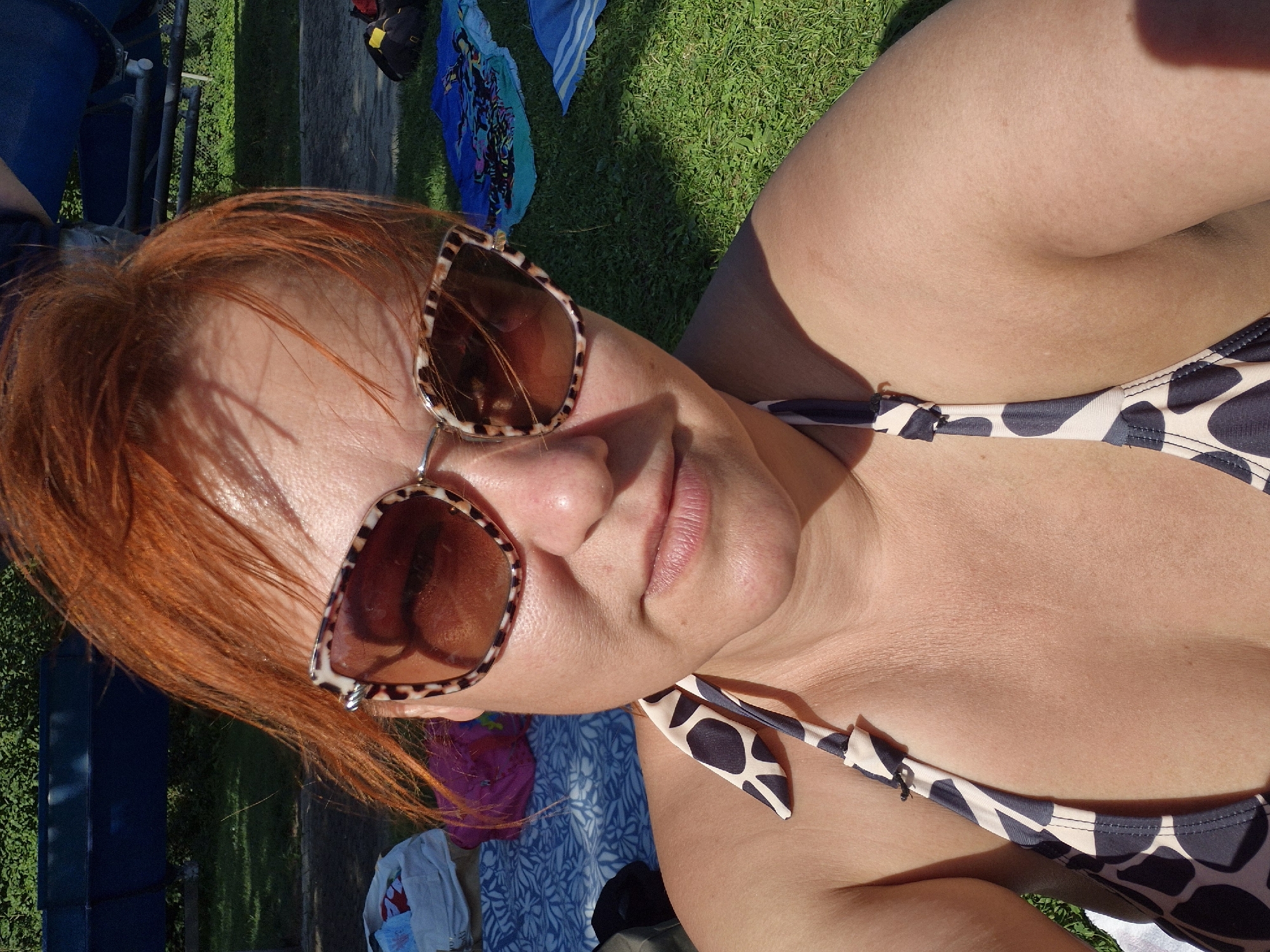 This is a close-up, outdoor selfie of a woman with bright red hair and large, leopard-print sunglasses. She is wearing a bikini top with a black and white giraffe-like pattern. The background suggests a sunny day at a recreational area, with green grass, a blue tarp or slide structure, and scattered towels or bags. Based on the provided location, this photo was likely taken in Schwäbisch Gmünd, Germany.