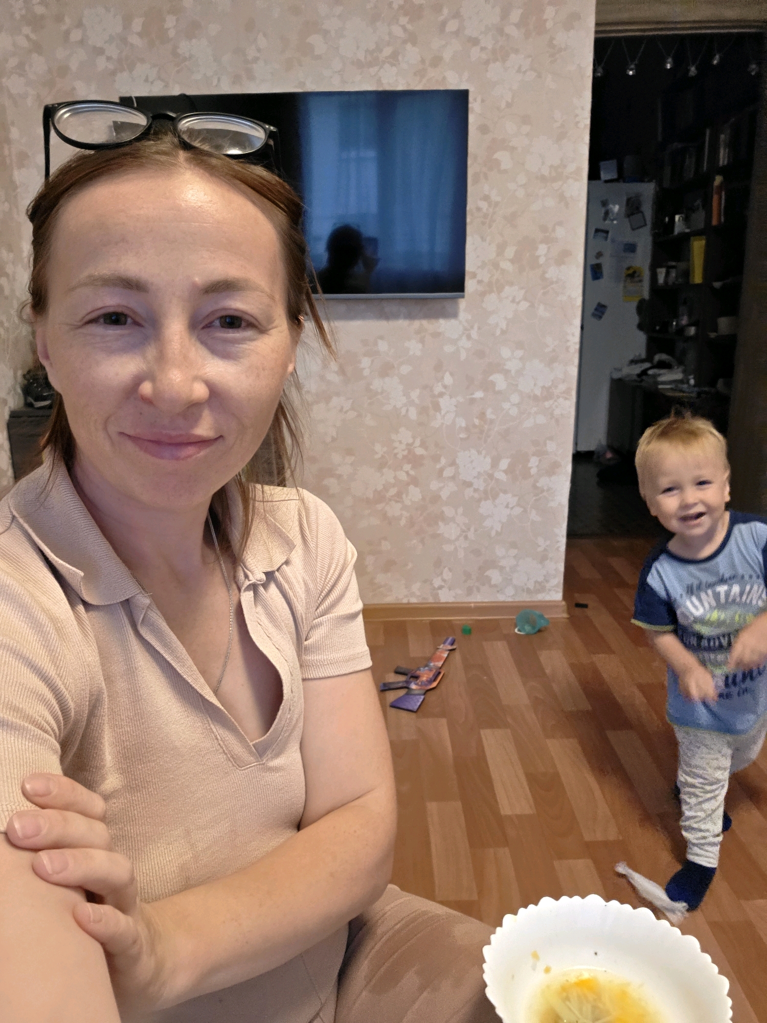 A woman is posing for a selfie in her apartment, with a young child visible in the background. The woman is wearing glasses on top of her head and a light-colored collared shirt. She has her arm crossed and is smiling at the camera. The child is wearing a blue and white t-shirt and light-colored pants, and appears to be looking towards the woman or camera. The room has light-colored floral wallpaper and a wooden floor. There are toys on the floor, including a toy gun. In the background, a television is mounted on the wall and a doorway leads to another room with shelves and a refrigerator. The scene is consistent with an indoor residential environment, likely in a city apartment.