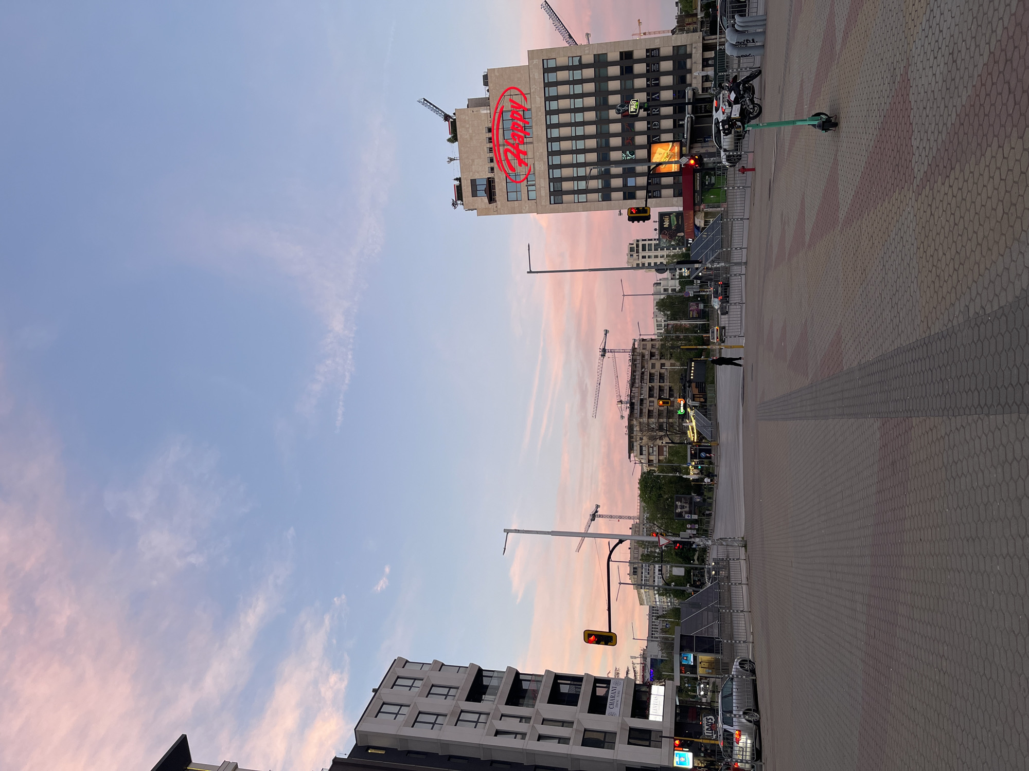 The image shows a cityscape at dusk in Sofia, Bulgaria. A tall building with the illuminated "Adler" logo on top dominates the scene. Construction cranes are visible in the background. Streetlights and traffic signals are present. There are buildings on each side of the street, and a wide sidewalk composed of interlocking hexagonal pavers. An electric scooter is parked on the sidewalk. There is a person walking in the street in the middle of the frame. The sky is filled with clouds that have a pinkish hue due to the sunset.