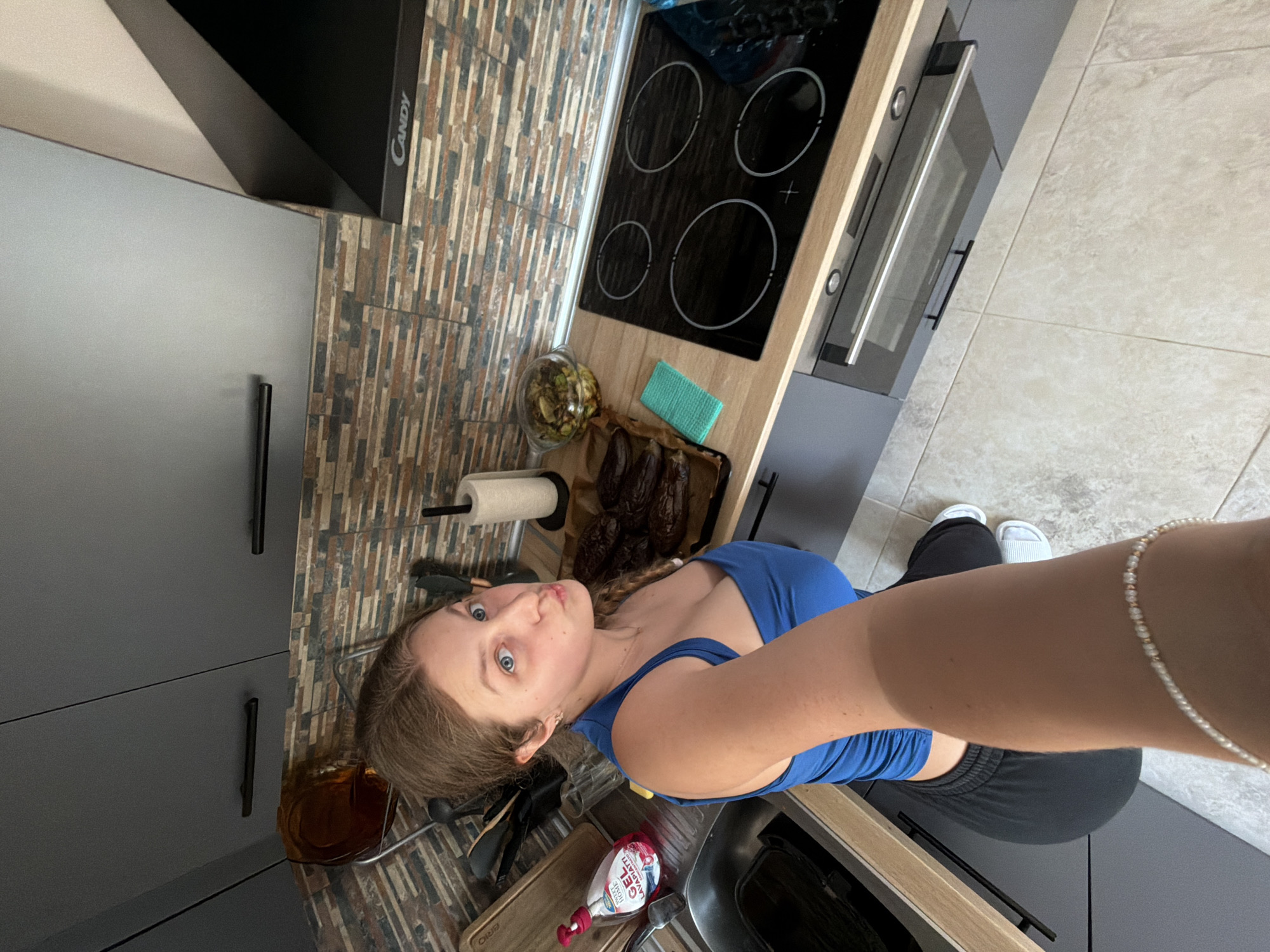 A woman takes a selfie in a modern kitchen with gray cabinets, a cooktop, and an oven. A dish of food and a roll of paper towels are on the counter. She is wearing a blue top and black pants. The floor is tiled. Location: possibly Litakovo, Bulgaria, based on inference, but not explicitly identifiable in the image.