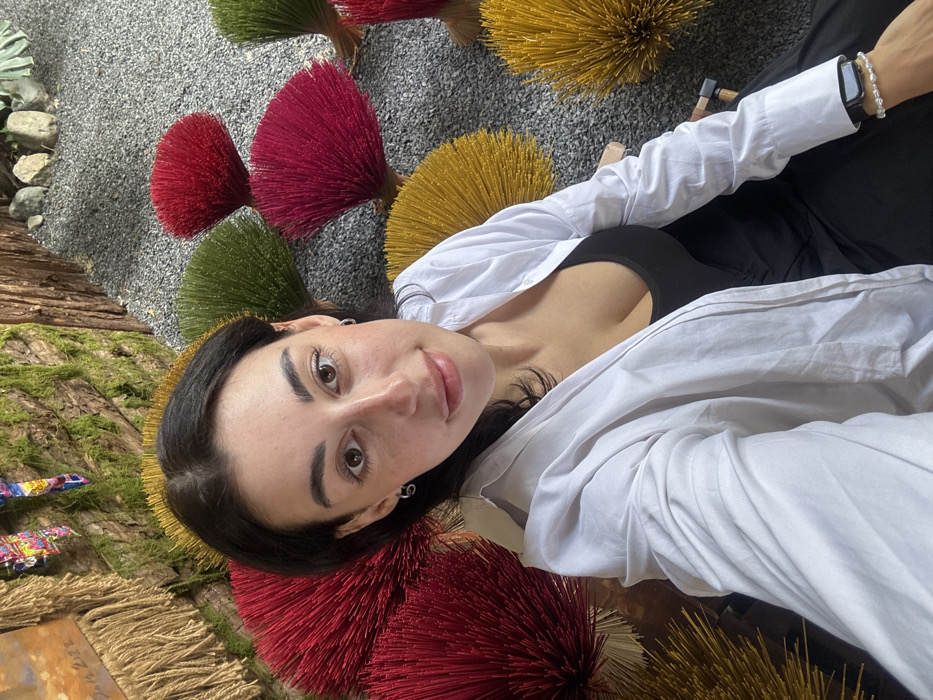A woman lies on her side amid colorful circular displays of red, green, and yellow straw-like material. She wears a white button-down shirt over a black top and dark pants. Her face is visible, with dark hair pulled back and light skin. A smartwatch with a pearl bracelet is on her wrist. The background features gray gravel and a wooden structure covered in moss and other decorative elements.