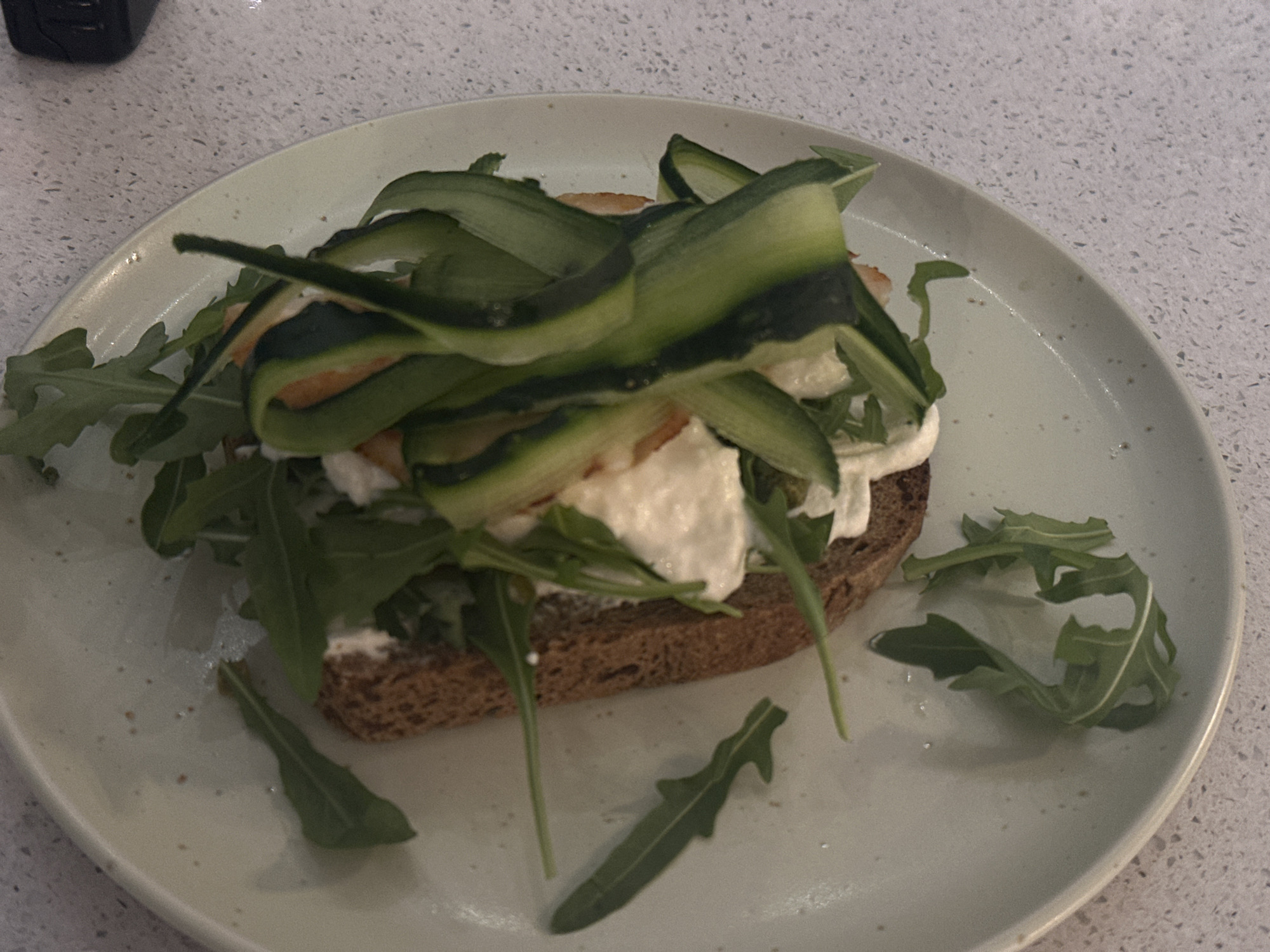 A close-up shot displays a plate of food, seemingly a sandwich or open-faced tartine, resting on a light-colored countertop with subtle speckles. The sandwich is composed of a slice of dark bread, topped with what appears to be soft cheese, arugula, and ribbons of green vegetable, possibly zucchini. Some arugula leaves are scattered around the sandwich on the plate. The overall impression is one of a simple, fresh, and possibly homemade dish. There is nothing to indicate that the image was taken in Novi Sad, Serbia.