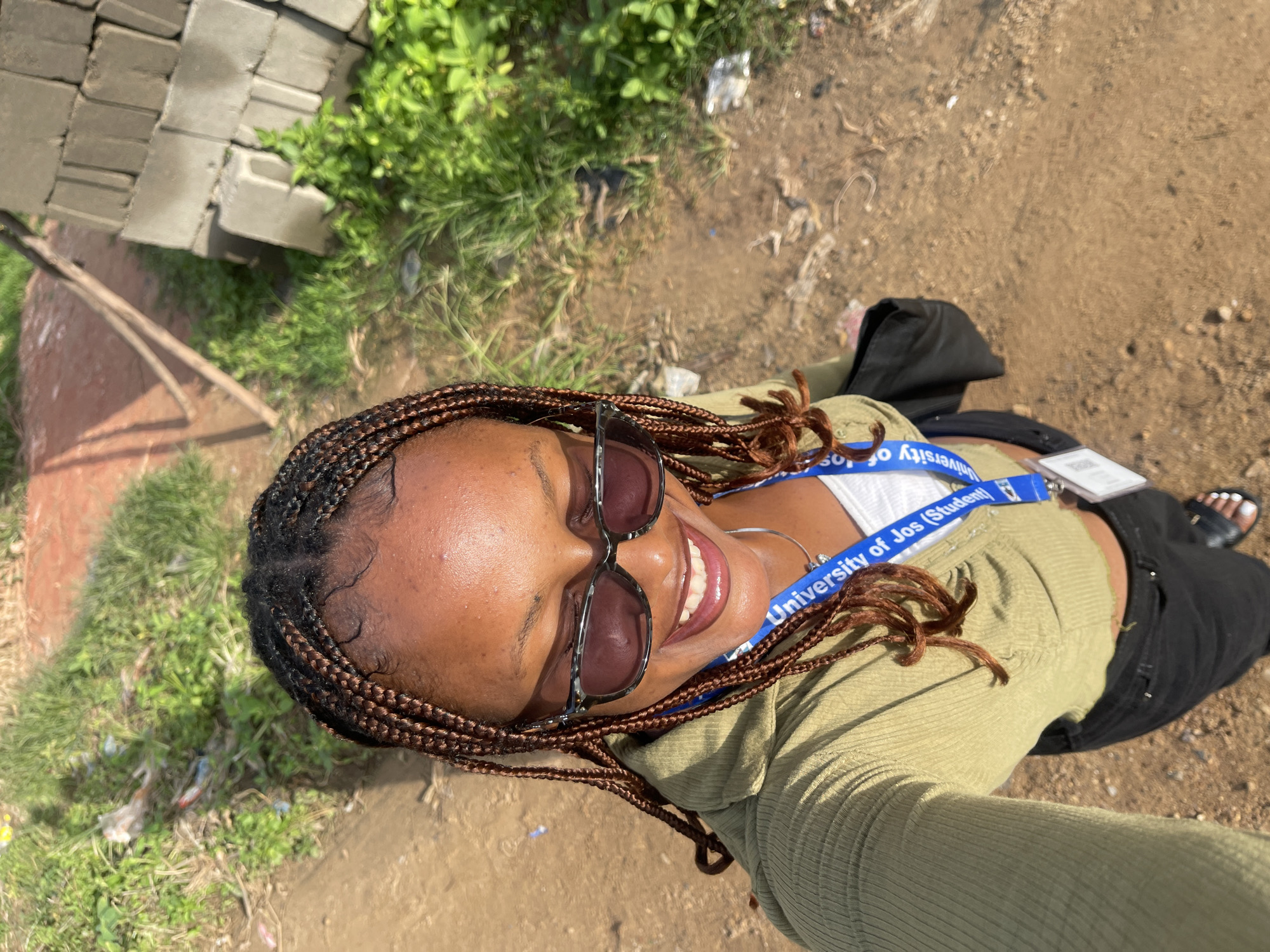 A medium shot captures a Black woman from the chest up, taking a selfie outdoors in what appears to be Rafin Par, Nigeria. She wears a green, ribbed, cropped long-sleeved shirt over a white tank top, black jeans, and black sandals. Her hair is styled in numerous brown braids that frame her face, and she wears dark, rectangular sunglasses. A blue lanyard with "University of Jos (Student)" printed on it hangs around her neck. She is smiling broadly, looking up and to her right. Behind her, the ground is dusty brown, with scattered debris and green grass to the left and right. To the far right, a stack of grey cinder blocks is visible, suggesting a construction or unfinished building site. The lighting indicates it is daytime, with shadows cast to the left.
