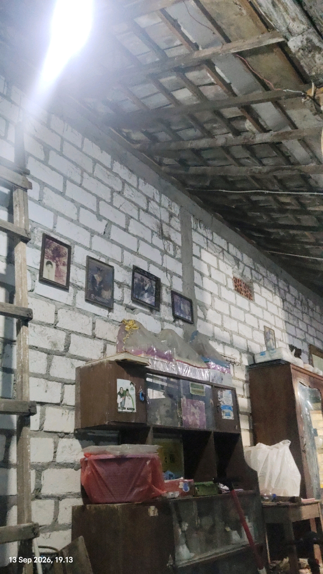 The image depicts the interior of what appears to be a storage area or a room under construction, located in Rengel, Indonesia, as indicated by the timestamp. The walls are constructed of white cinder blocks, with a rough, unfinished texture. The ceiling is exposed, showing a wooden frame structure with tiled roofing.
On the left side of the wall, there are four framed photographs hung in a row. The first photograph from the left shows a portrait of two people. The subsequent three photographs are smaller and appear to contain group photos or other framed images. Below the photographs, a wooden shelf unit is visible, with several compartments. One compartment on the left contains a red plastic container, partially covered by clear plastic sheeting. Above this shelf unit, on top of a dark wooden box, are two small framed pictures, one of which has the word "LOSS" visible.
To the right of the shelf unit, a large dark wooden cabinet or wardrobe is present. A white plastic bag is placed on top of a surface next to this cabinet. The floor area is cluttered with various objects, including what looks like a red broom handle. The overall impression is one of a functional but somewhat disorganized space. The lighting in the scene is a combination of natural light coming from the upper left, creating a bright glare, and ambient interior light, which casts shadows.