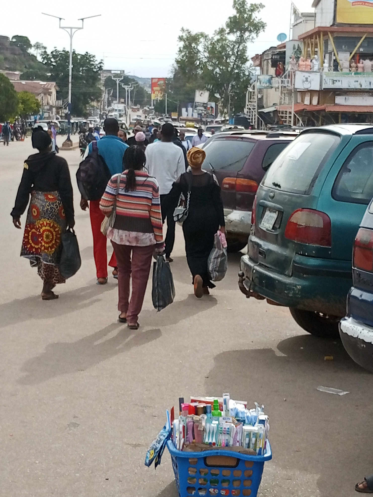 The image depicts a street scene in Jos, Nigeria, likely a commercial area given the presence of shops and a busy road. In the foreground, a blue plastic basket filled with toiletries, including toothbrushes and toothpaste, sits on the ground. Several people are walking on the street, some carrying shopping bags. A green station wagon is parked on the right side of the road, partially obscuring the view of other vehicles and the surrounding buildings. The overall atmosphere suggests a typical day in a bustling urban environment. The people in the image are diverse in age and gender, and their clothing suggests a casual style. The architecture of the buildings in the background appears to be a mix of modern and traditional styles, which is common in many Nigerian cities. The presence of street vendors and the variety of vehicles indicate a lively marketplace.