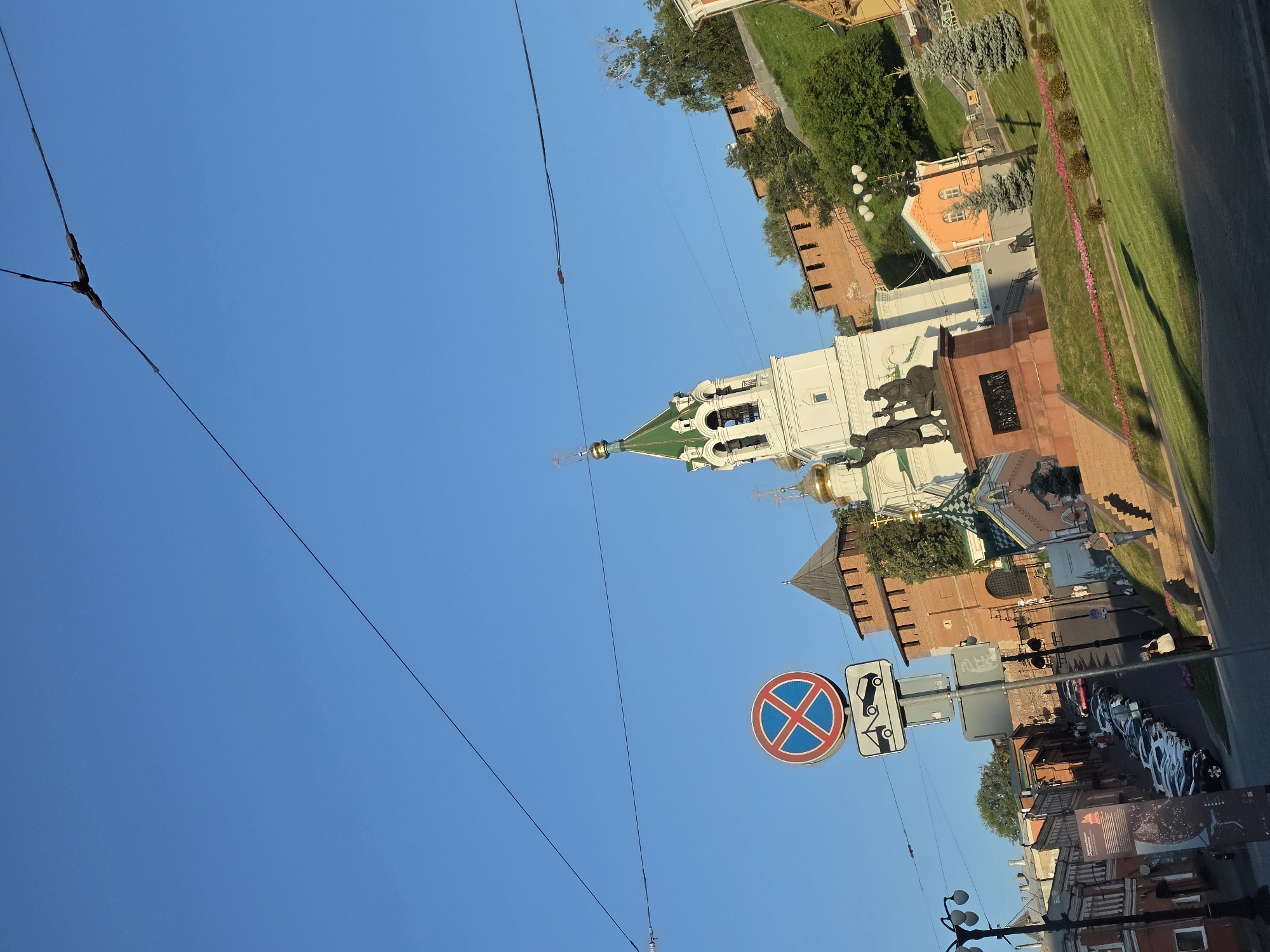 The image depicts a view of Nizhny Novgorod, Russia, featuring architectural landmarks under a clear blue sky. In the foreground, a prominent no parking sign with a tow-away symbol is visible, positioned near a street with parked cars. Adjacent to the sign is a sculpture of two figures on a pedestal. Behind the sculpture, a white bell tower with a green roof and golden domes rises, indicating a church. To the left of the bell tower, a portion of a red brick fortress or wall is visible, a characteristic feature of Nizhny Novgorod's Kremlin. The background shows more of the red brick fortifications, lush green trees, and a small orange building. The scene suggests a bright, sunny day.