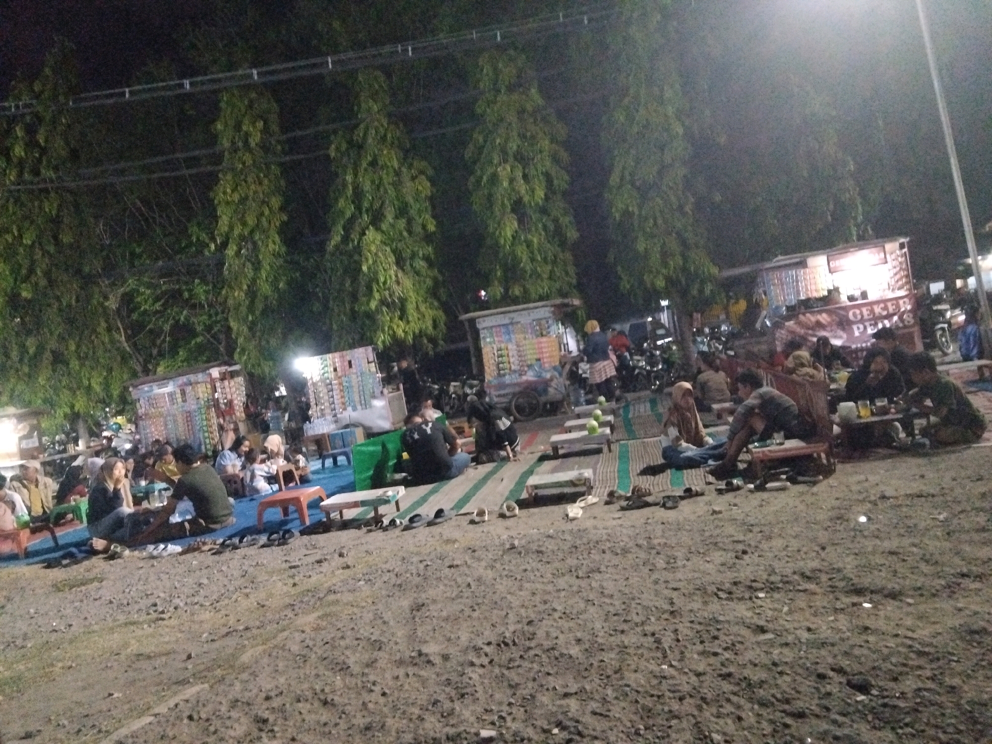 The image depicts an outdoor gathering scene at night, likely a food stall or market, located in Sedati Agung, Indonesia. Several people are seated on mats on the ground, some at low tables, while others are standing. Food stalls are visible, with one clearly displaying "GEKER PEDAS" signage, suggesting a food vendor. Motorcycles are parked in the background, indicating a public gathering place. The environment is a mix of gravel and matting, with trees lining the background, partially illuminated by artificial light. The overall atmosphere is informal and social, with people enjoying food and conversation in a casual setting.