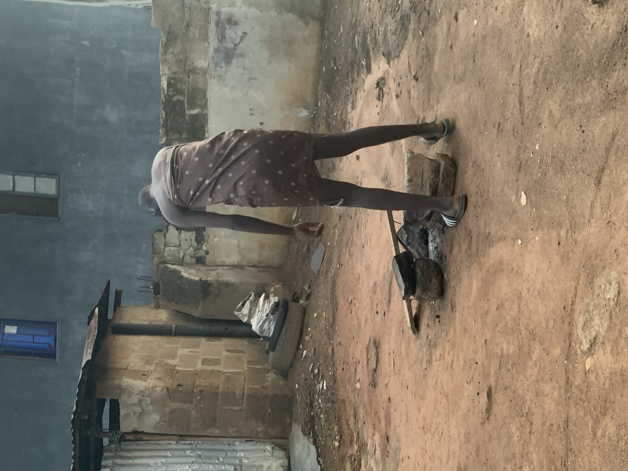 In Shagamu, Nigeria, a person wearing a brown cloth patterned with white polka dots stands in a dusty, outdoor area. They are facing away from the camera, with their head lowered. The person is wearing flip-flops. In front of them, on the dirt ground, is a makeshift cooking setup with rocks and a piece of wood. To the left, there is a building constructed with large, rough stones and a corrugated metal roof. A black pipe extends upwards from near the building. Further left, a section of corrugated metal siding is visible. Behind the person, a light blue wall with two windows is the backdrop, with more stone structures and rough walls to the sides. The ground is covered in reddish-brown dirt with scattered debris.