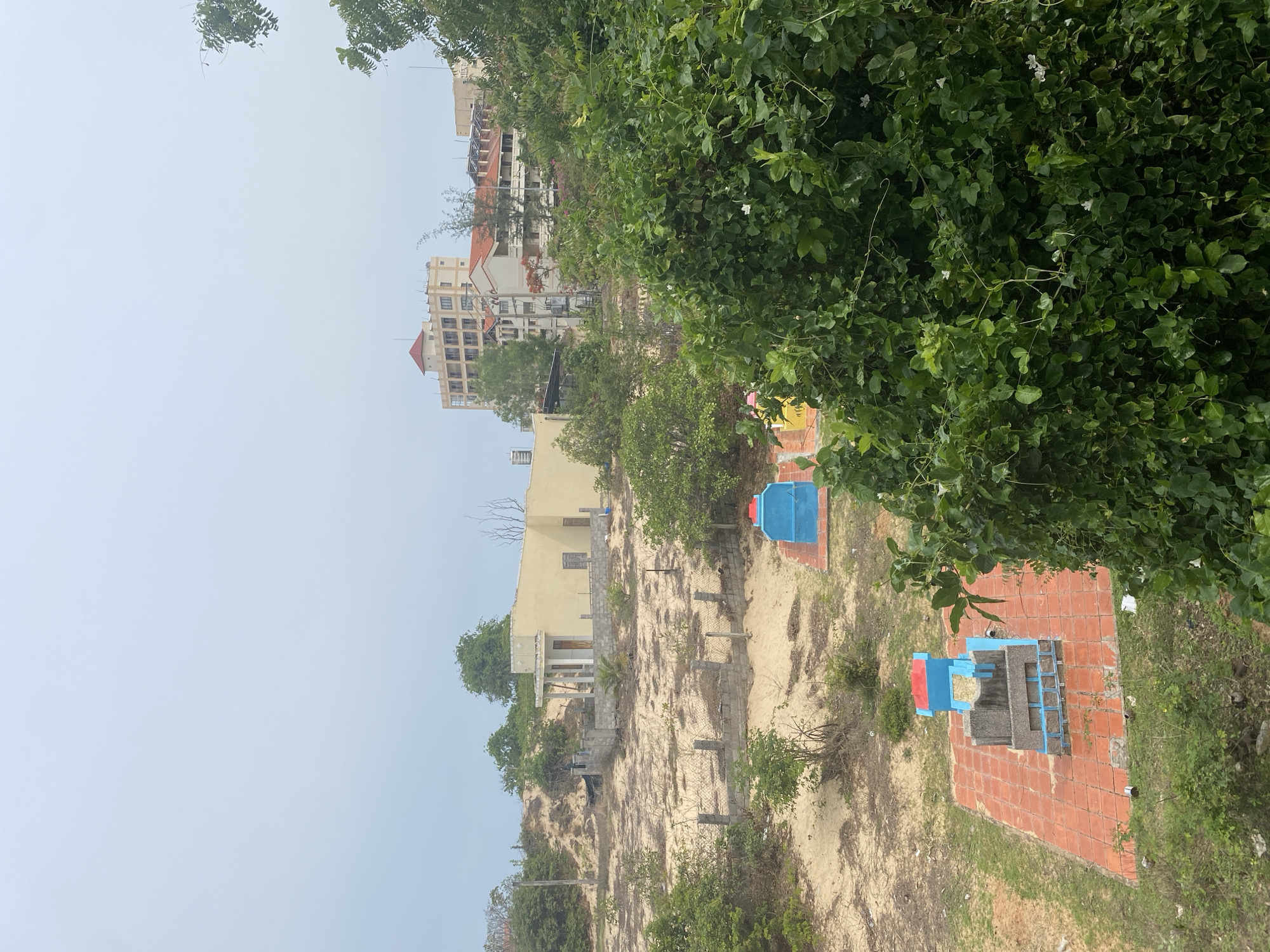 The image depicts a view in Mũi Né, Vietnam, showcasing a landscape transitioning from urban development to more natural terrain. In the background, there are multistory buildings with light-colored facades and red-tiled roofs, suggesting a mix of residential and commercial structures. Further down the hillside, there is a pale yellow building, possibly a residence or smaller establishment, situated amidst a sandy, undeveloped area. The ground is sandy with sparse vegetation. In the foreground, there is a tiled area with painted blue and red concrete structures, possibly part of a small park or recreational space. Dense green foliage occupies the right side of the image, framing the scene. The sky is overcast, and the angle of the image suggests it was taken from a slightly elevated position.