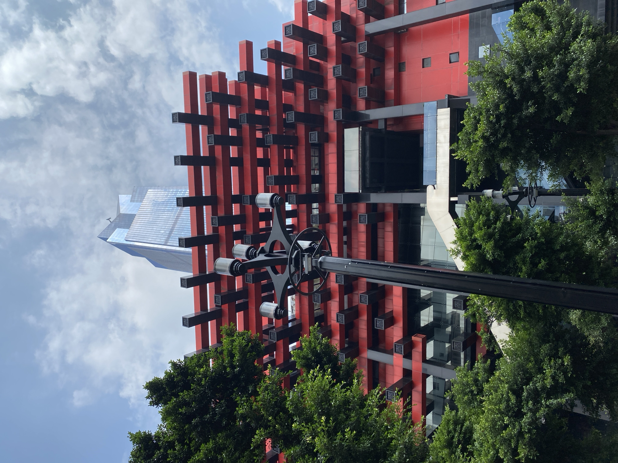 The image shows the Chongqing Grand Theatre, located in the Yuzhong District of Chongqing, China. The theatre is characterized by its striking red facade adorned with a grid of black beams, creating a layered effect. In the foreground, lush green trees partially obscure the lower levels of the building. A black lamppost with multiple light fixtures stands prominently in the center of the frame, adding a modern element to the scene. Behind the theatre, a contemporary skyscraper with a reflective glass exterior rises into the sky. The sky is partly cloudy, with white clouds scattered across a blue expanse. The perspective is from a low angle, looking upwards at the architecture. There are no people visible in the image.