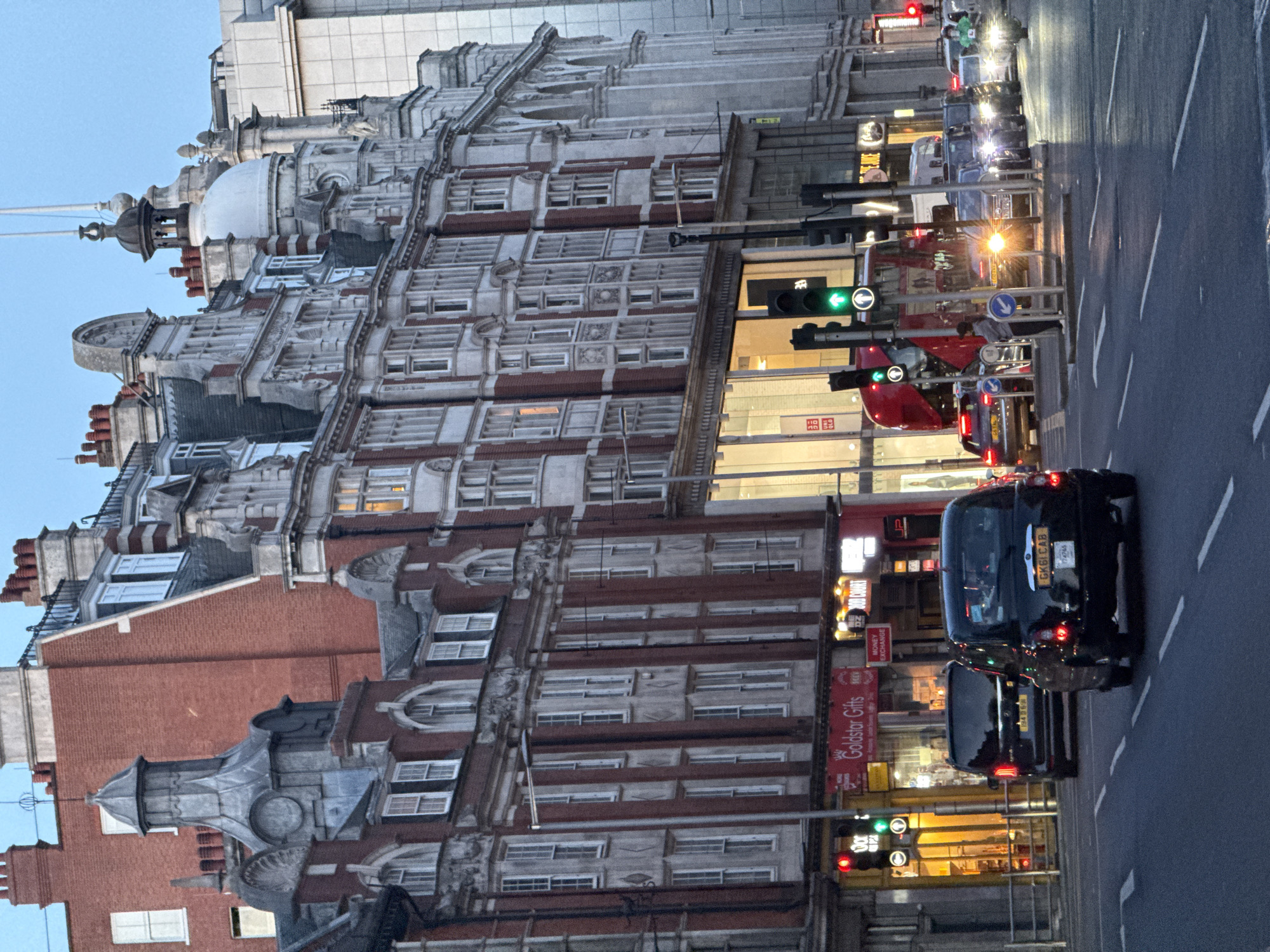 The image shows a street scene in London, United Kingdom. The architecture is notable, with a large, ornate building occupying a significant portion of the frame. The building has a primarily white facade with red brick accents. Several domes and architectural details adorn the top of the building. A multilane road runs adjacent to the building, with vehicles traveling in both directions. Traffic signals are present, and at least one of the lights is green. There is a double decker red bus, a red car and several black taxis on the road. The image was likely taken in the evening, given the light conditions and the illumination of the building's windows.