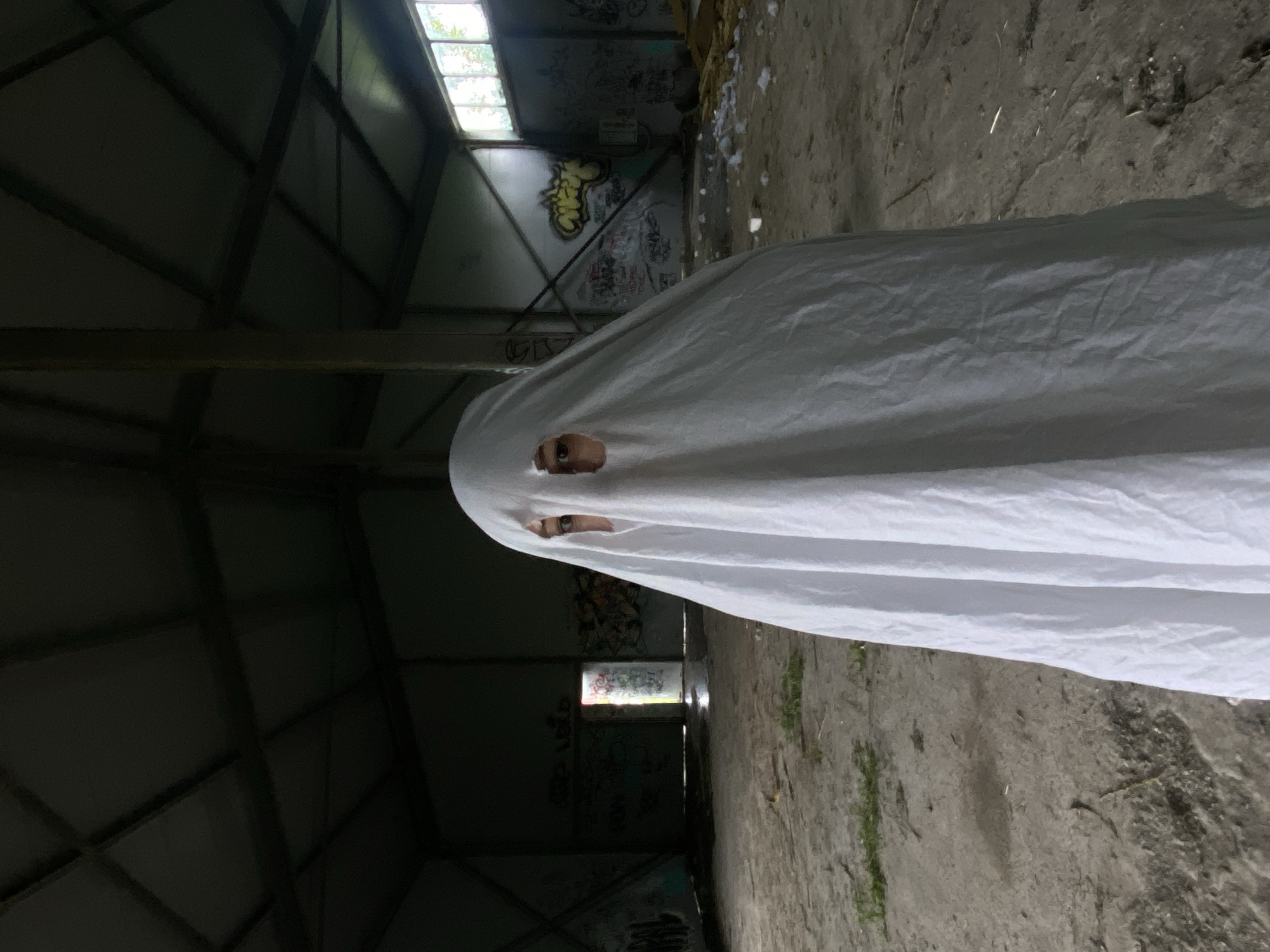 A person, draped in a white sheet to resemble a ghost, stands in the center of an abandoned industrial building in Khabarovsk, Russia. The building's interior is dimly lit, with a corrugated metal roof and exposed beams. The concrete floor is dusty and uneven, with patches of sparse vegetation. Graffiti covers the walls, and debris is scattered around the space. Two windows, illuminated by daylight, are visible in the background. The person's eyes are visible through holes cut in the sheet.