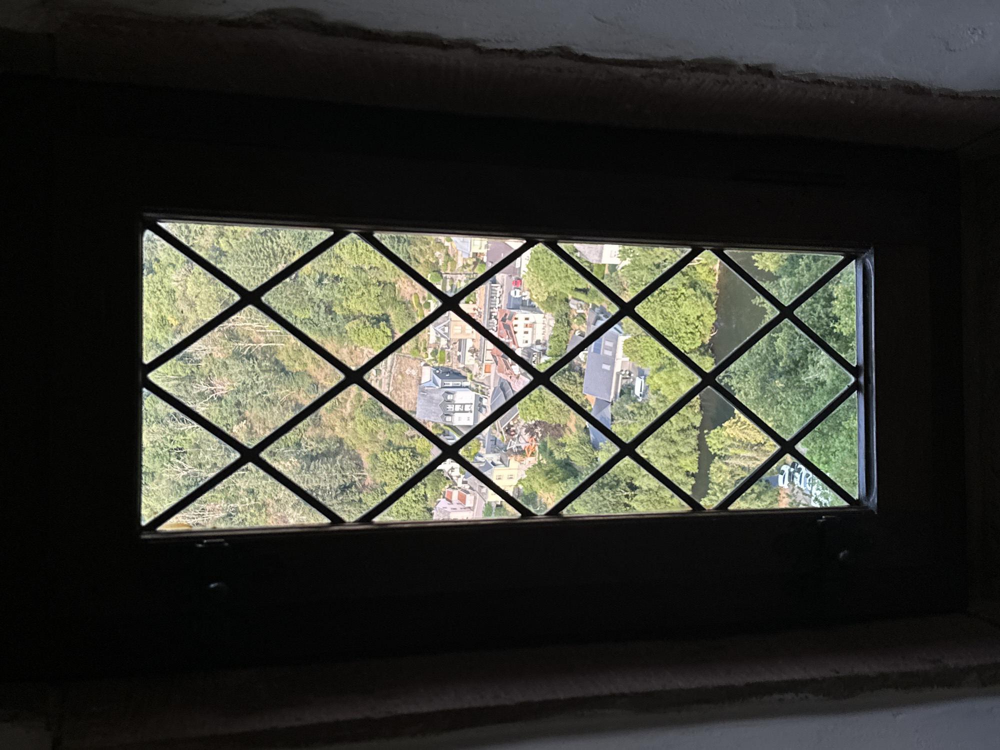 A vertical shot shows a view through a leaded glass window overlooking a town nestled in a valley. The window is divided into diamond-shaped panes by dark mullions. The view outside reveals a dense forest on a hillside that slopes down towards a river. A small town with red-tiled roofs and lighter-colored buildings is situated between the forest and the river. Some of the buildings appear to be traditional European architecture. A few vehicles are visible on a road winding through the town. The image is taken from a darkened interior, with the window frame and surrounding wall providing a stark contrast to the brightly lit outdoor scene.