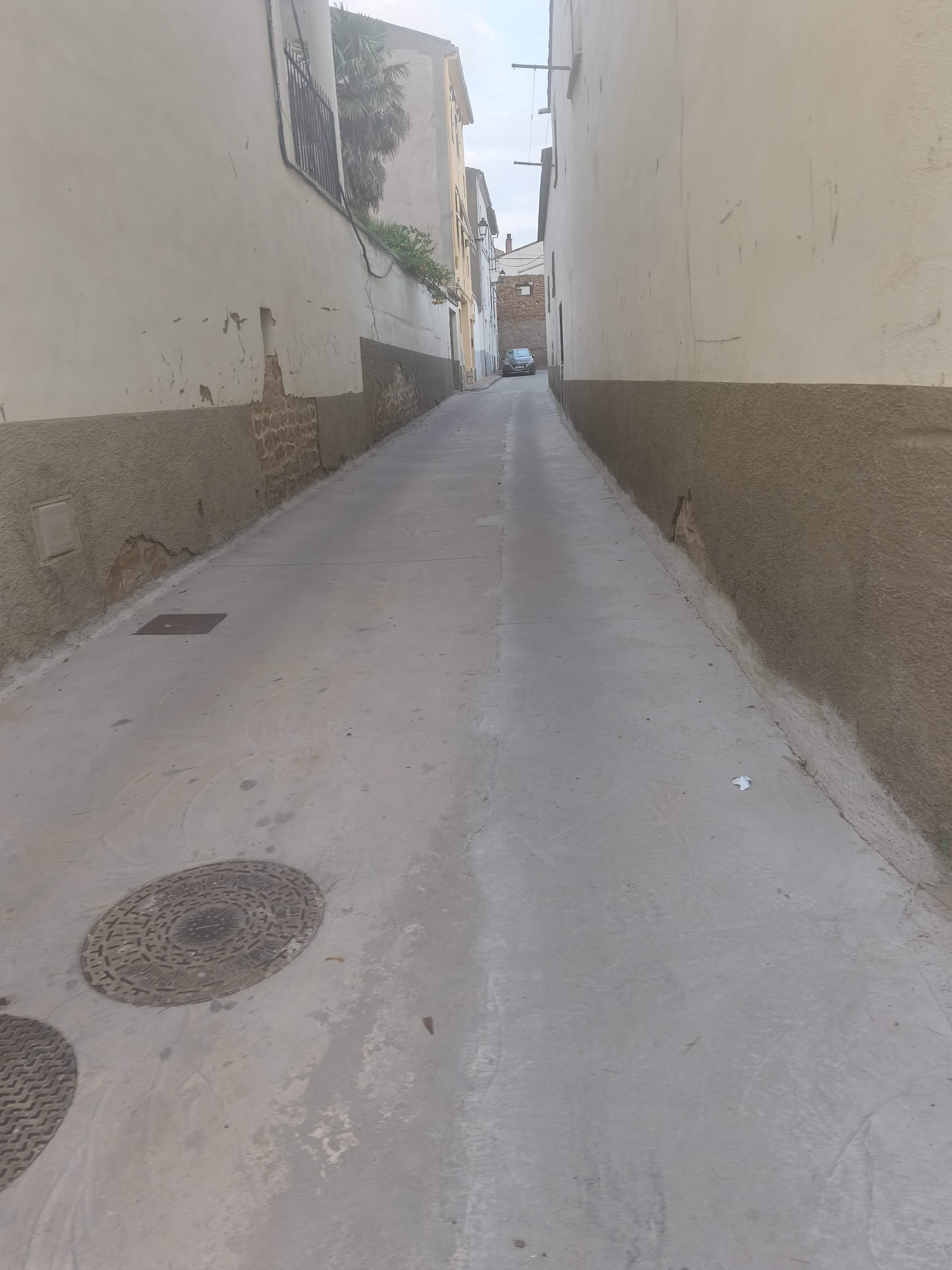 The image depicts a narrow, unpaved alleyway in the town of Híjar, Spain, with buildings lining both sides. The walls of the buildings are made of light-colored stucco and feature exposed brick sections near the base, indicating age and some disrepair. One building on the left has a dark metal railing and a patch of green vegetation. On the right, the wall is a solid brown stucco. The ground of the alley is a smooth, light gray concrete surface, marked by some water stains and what appear to be tire tracks. Two circular manhole covers are visible on the ground, one to the left and another further down the alley. In the distance, a small, dark-colored car is parked. The sky is visible at the top of the frame, appearing light and overcast. The overall impression is of a quiet, residential street with a somewhat weathered appearance.