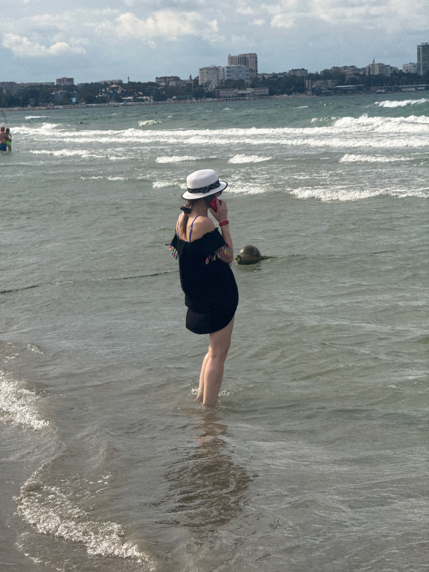 A woman stands in the shallow water of the Black Sea in Anapa, Russia, facing away from the camera and speaking on a mobile phone. She is wearing a white hat with a black band, an off-the-shoulder black dress adorned with colorful tassels, and a red bracelet on her left wrist. Her legs are visible from the knees down, submerged in the water. In the background, a coastline with buildings, including apartment blocks and what appears to be a Ferris wheel, is visible under a cloudy sky. The sea is choppy with white-capped waves rolling towards the shore. Two people are visible in the distance, standing in the water further out.