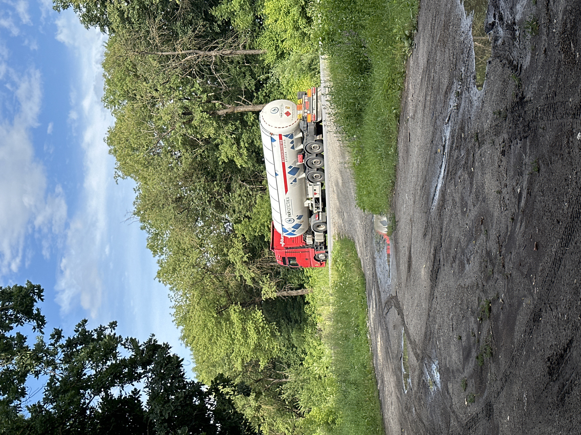 A red semi-truck with a white tanker trailer is driving on a paved road that curves to the right. The truck has "BDUM" written on its side in white text, and the trailer has "UKRLOGISTICS" written on it in white text with a blue logo. The truck is surrounded by lush green trees and foliage on both sides of the road, with a bright blue sky and white clouds above. In the foreground, a dirt path with tire tracks and several puddles of water is visible. The overall scene suggests a rural or semi-rural setting with a clear day.