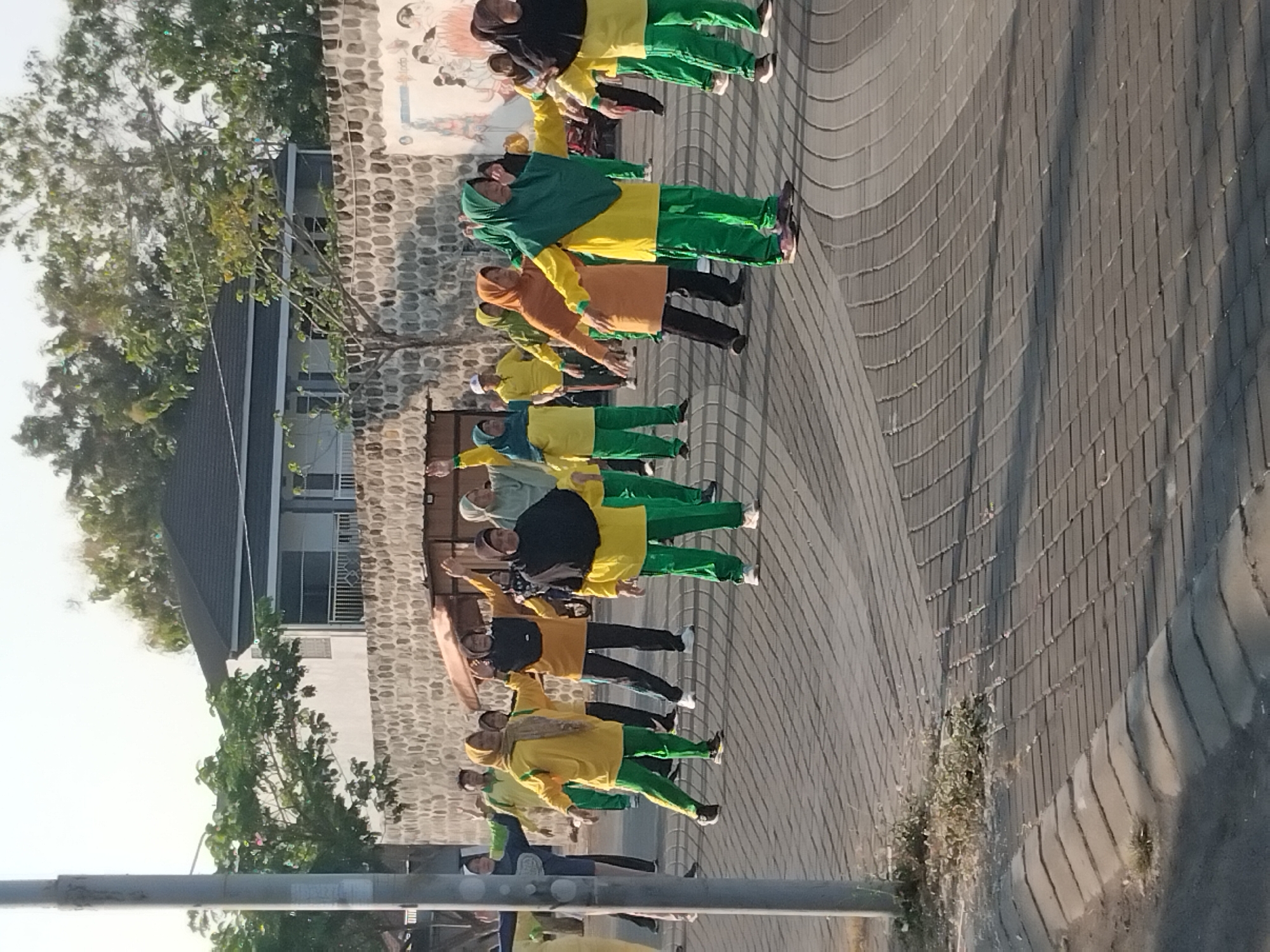 In Karanganyar, Indonesia, a group of people, primarily women wearing hijabs and exercise attire in shades of yellow, green, and orange, are engaged in an outdoor aerobic exercise session on a paved surface. The background features a stone wall with a mural on the right side and a building with a dark roof. The scene is illuminated by natural sunlight, casting shadows on the ground. A lamppost is visible on the left side of the image. The terrain is a street or plaza paved with interlocking bricks, with a curb and sidewalk area on the lower left.
