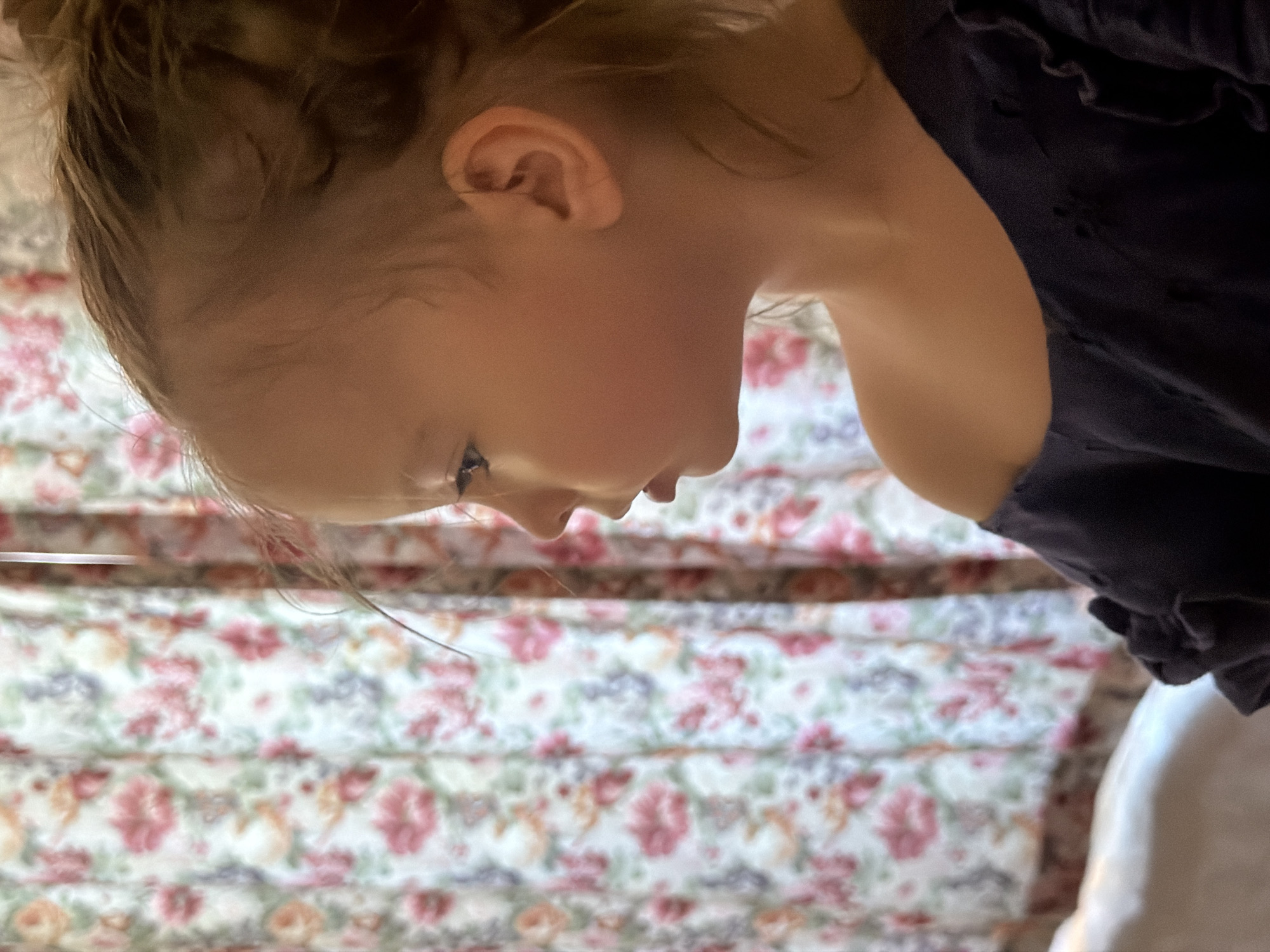 A young girl is pictured in a close-up, profile view, facing left. She has fair skin and her hair is pulled back. Her eyes are open and she is looking towards something out of frame. She is wearing a dark-colored, off-the-shoulder top. In the background, floral curtains are visible, with a pattern of pink, white, and green flowers against a lighter background. The lighting suggests an indoor setting, possibly near a window. The scene is static and shows a moment of quiet observation. This image was likely captured in Fethiye, Turkey, given the context provided.