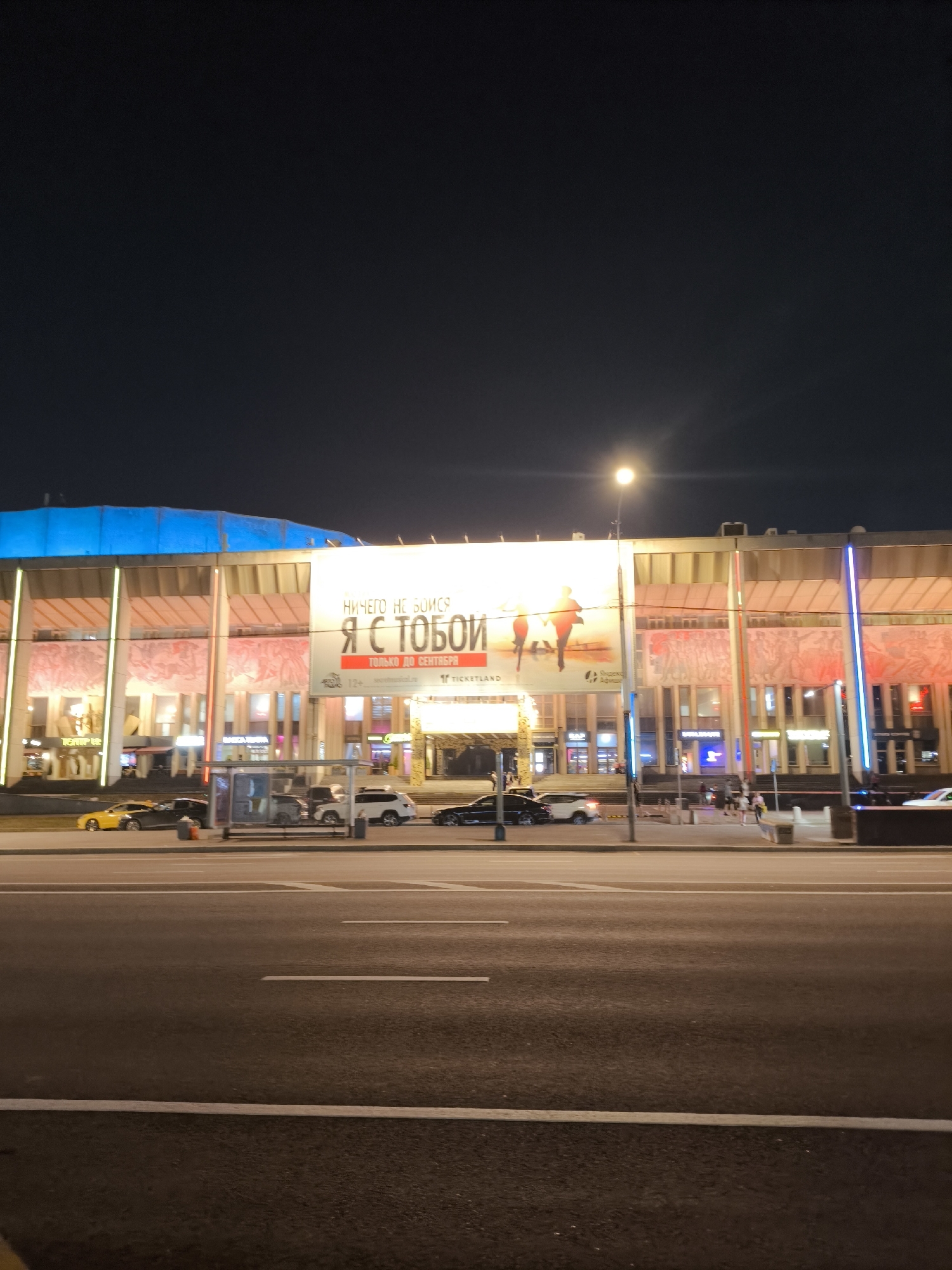 The image shows a wide shot of the facade of a large building at night, likely a cultural or entertainment venue, in Moscow, Russia. A large illuminated billboard is prominently displayed on the building's facade, featuring text in Russian which appears to be an advertisement. The billboard includes silhouettes of two figures. The building itself has a modern architectural style with large windows and illuminated columns. In front of the building, there is a street with several cars parked and driving, and a bus stop shelter. The overall atmosphere is urban and active, with artificial lighting dominating the scene.