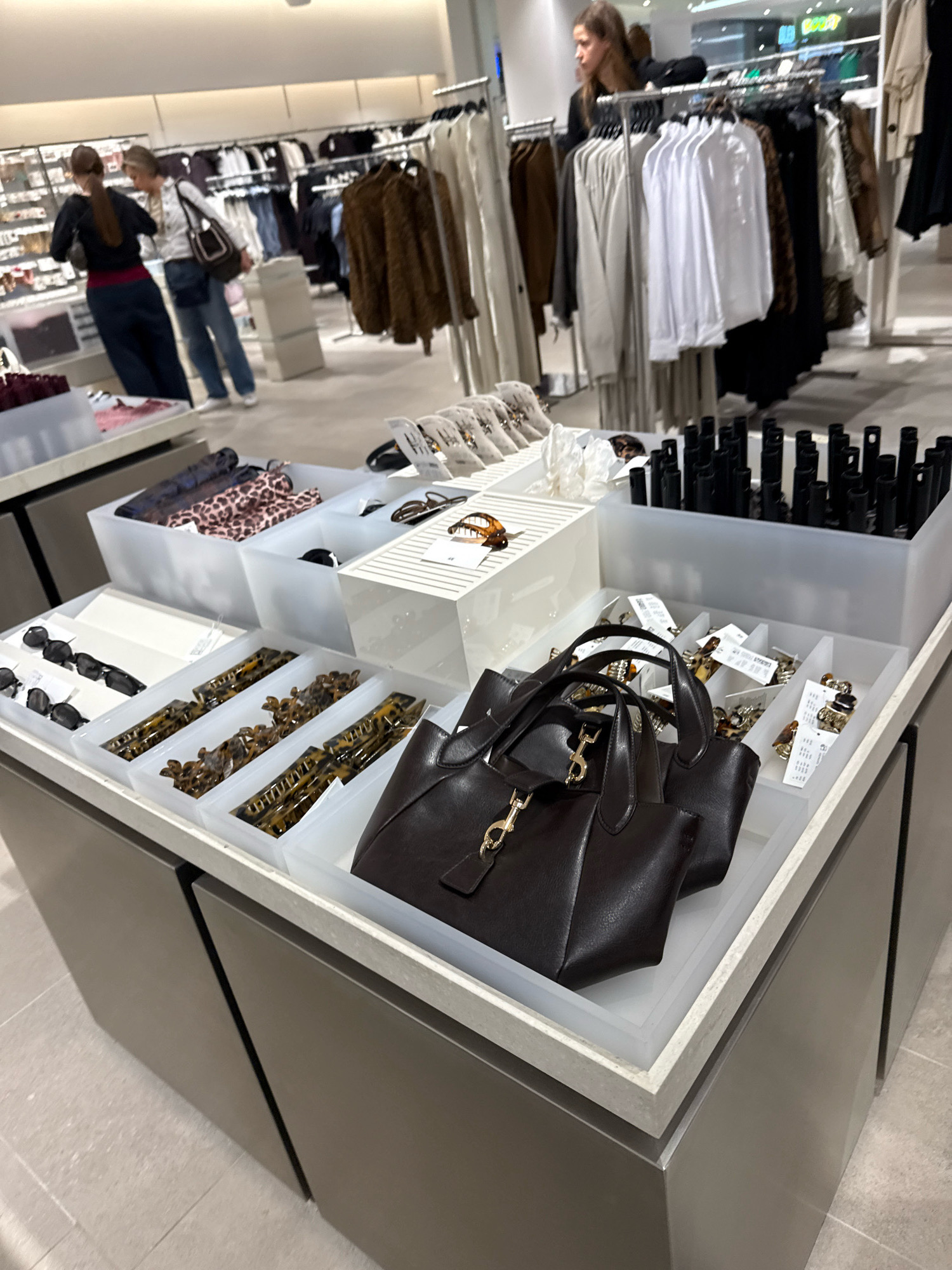 This image depicts an interior view of a retail clothing store, likely a department store or a fashion boutique, in Tallinn, Estonia. The foreground displays a section of a store counter with various accessories arranged in white trays. These accessories include several pairs of dark sunglasses, multiple hair claw clips with a tortoiseshell pattern, and a dark brown leather handbag with gold hardware. Behind the counter, a collection of black cylindrical items, possibly cosmetic products like lipstick or mascara, are displayed in a white tray. Further back, clothing racks are visible, stocked with a variety of garments, including blouses, jackets, and sweaters in neutral and patterned fabrics. Two women are partially visible in the mid-ground, one in the foreground wearing a dark top and burgundy pants, and another in the background wearing a white top and jeans, seemingly browsing the merchandise. The store appears to be well-lit with artificial lighting. The flooring consists of light-colored tiles. The overall impression is a well-organized retail environment offering fashion accessories and apparel.