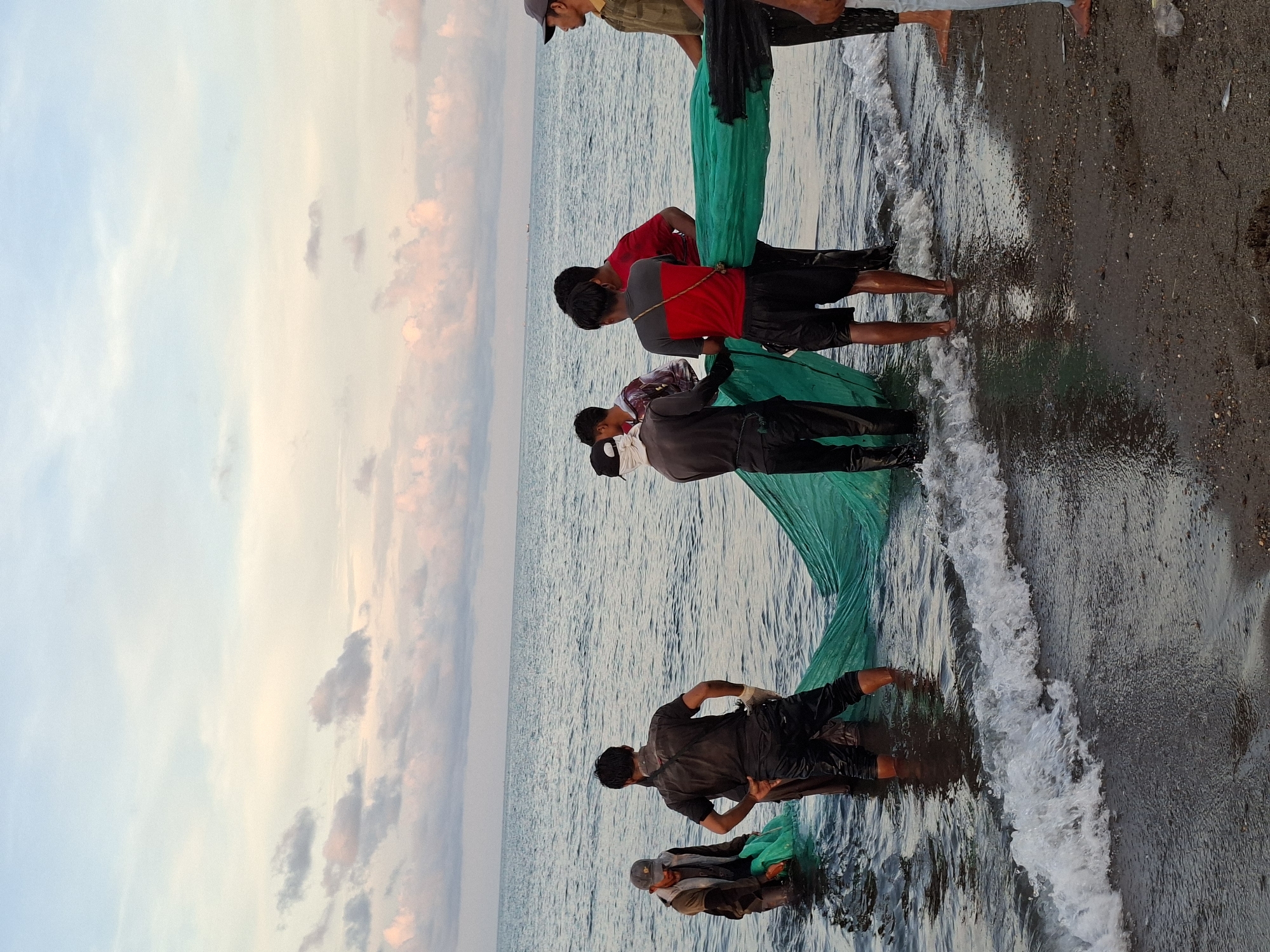 The image depicts a group of men fishing with a large green net in the surf along a beach. The location is likely near Lhokseumawe, Indonesia, based on the cultural characteristics of the people and the coastal environment. The men are standing in the shallow water, holding and pulling the net. The sky is visible in the background, featuring clouds tinged with pink and orange. The beach has dark sand. Waves are washing onto the shore.
