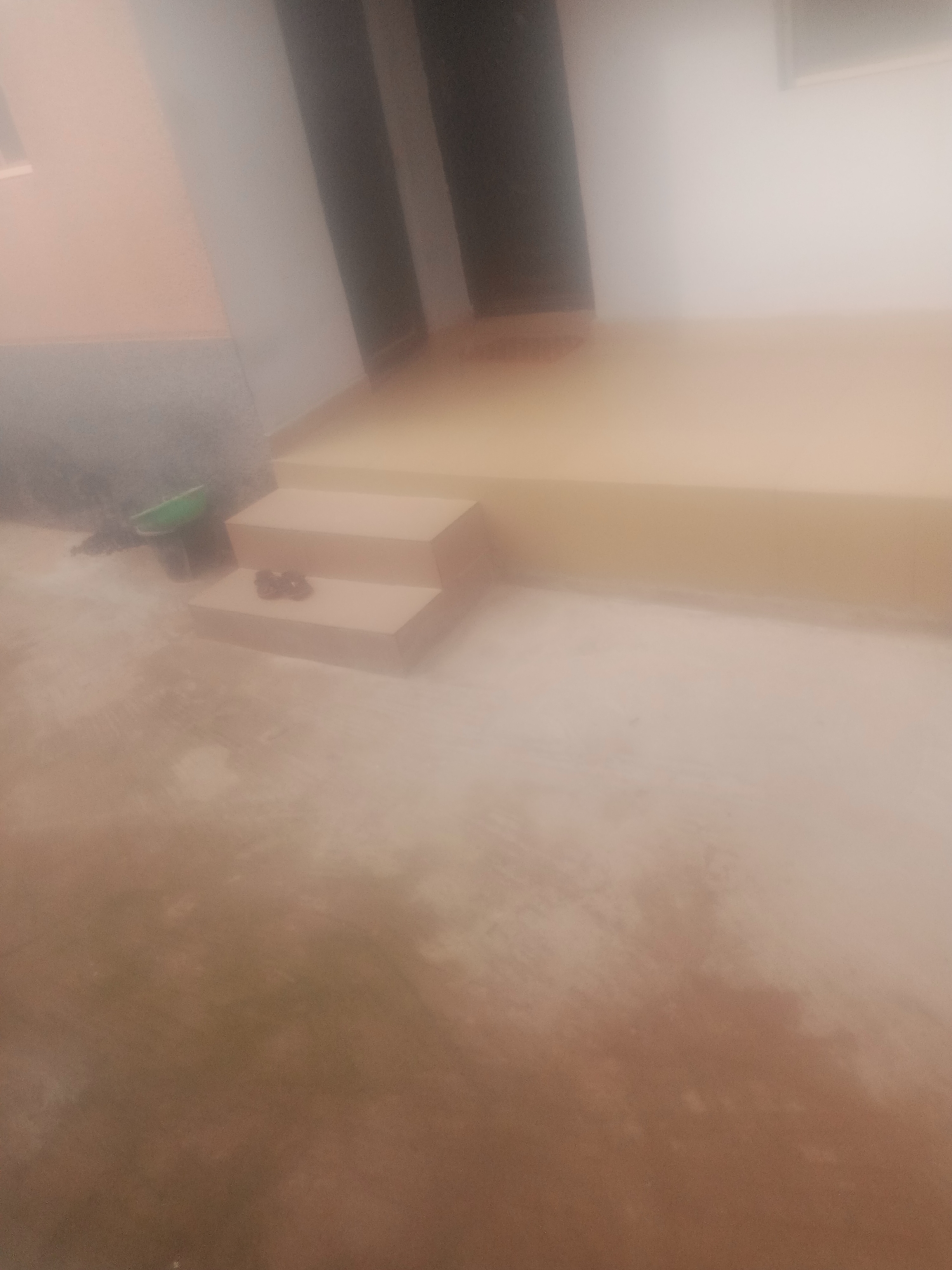 A blurry, outdoor scene shows a set of two concrete steps leading up to a light brown colored landing. A green shovel is resting on the ground near the bottom of the steps, and a pair of dark colored flip-flops are on the lower step. The ground appears to be a dirt or concrete surface. The background shows a light brown wall and a darker vertical structure.