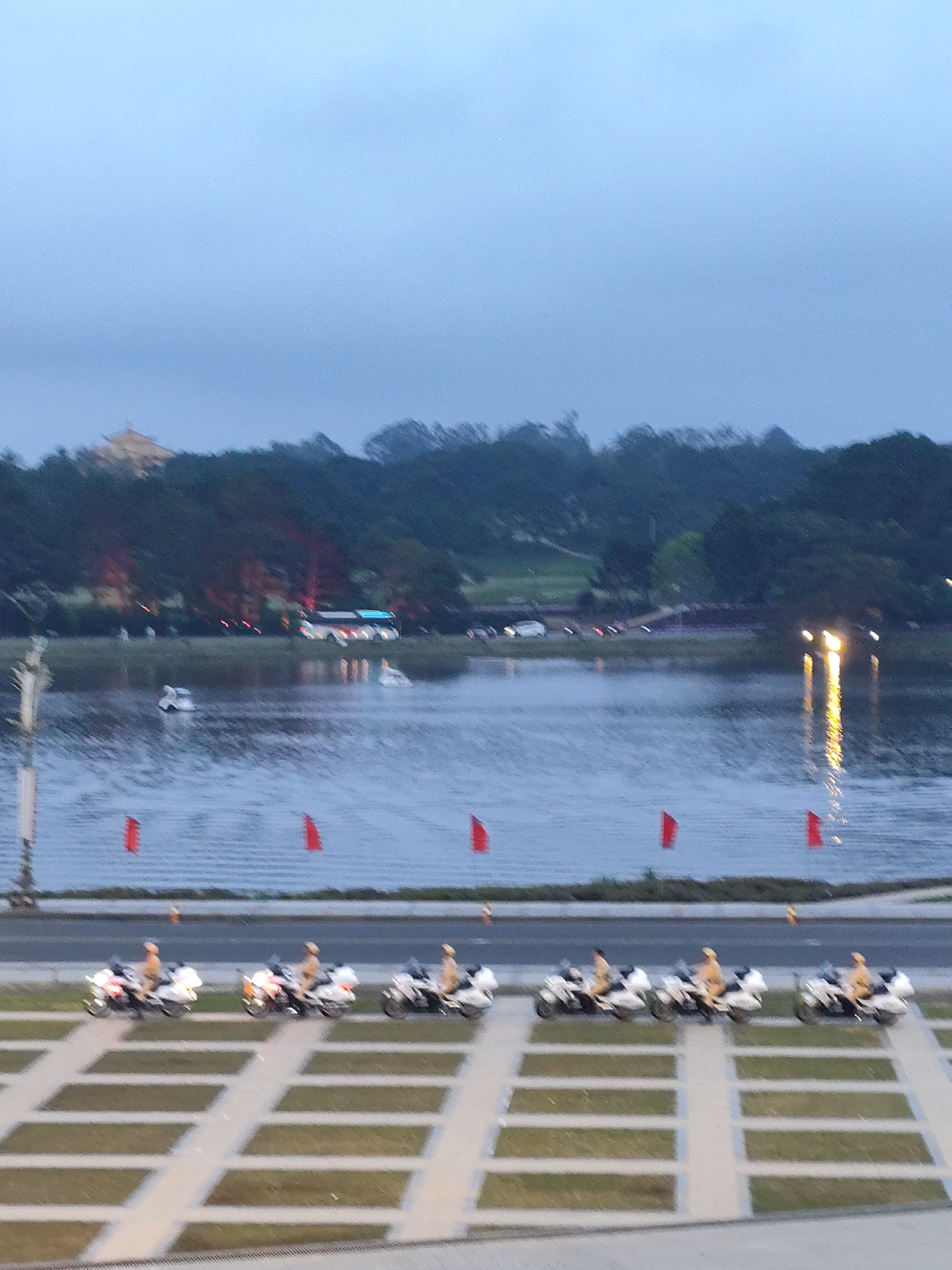 The image depicts Xuan Huong Lake in Da Lat, Vietnam, at dusk. A line of motorcycles with riders in uniform is parked along the lakeside promenade. The motorcycles are white with prominent windshields, and the riders are wearing light-colored uniforms. In the background, across the lake, a dense line of trees forms the horizon, with a few structures visible within them. A large bus with illuminated windows is seen driving along a road in the distance. The lake itself is calm, with reflections of the sky and lights visible. Several red flags are planted along the edge of the road. The overall atmosphere is serene and muted, characteristic of the late evening hours.