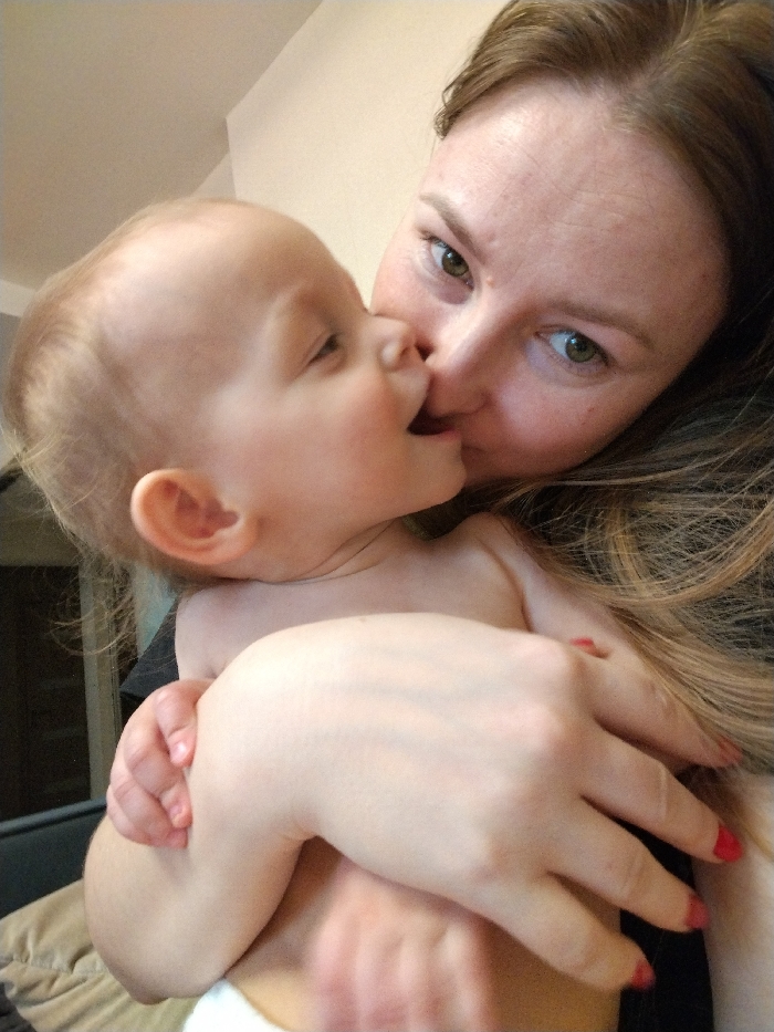 An adult female and an infant are depicted in a close-up, indoor setting. The adult female, with light brown hair and green eyes, looks directly at the camera. Her right hand, featuring red-painted fingernails, cradles the infant. The infant, with light hair, is positioned with its face close to the adult's, their noses nearly touching, and its mouth slightly open. The infant's left hand grips the adult's arm. The infant appears partially unclothed, revealing its bare torso. The background consists of a plain, light-colored wall, with a dark frame and a beige cushion visible in the lower left. The specific location within the city of Chaikovsky, Russia, cannot be determined from the visual evidence presented.