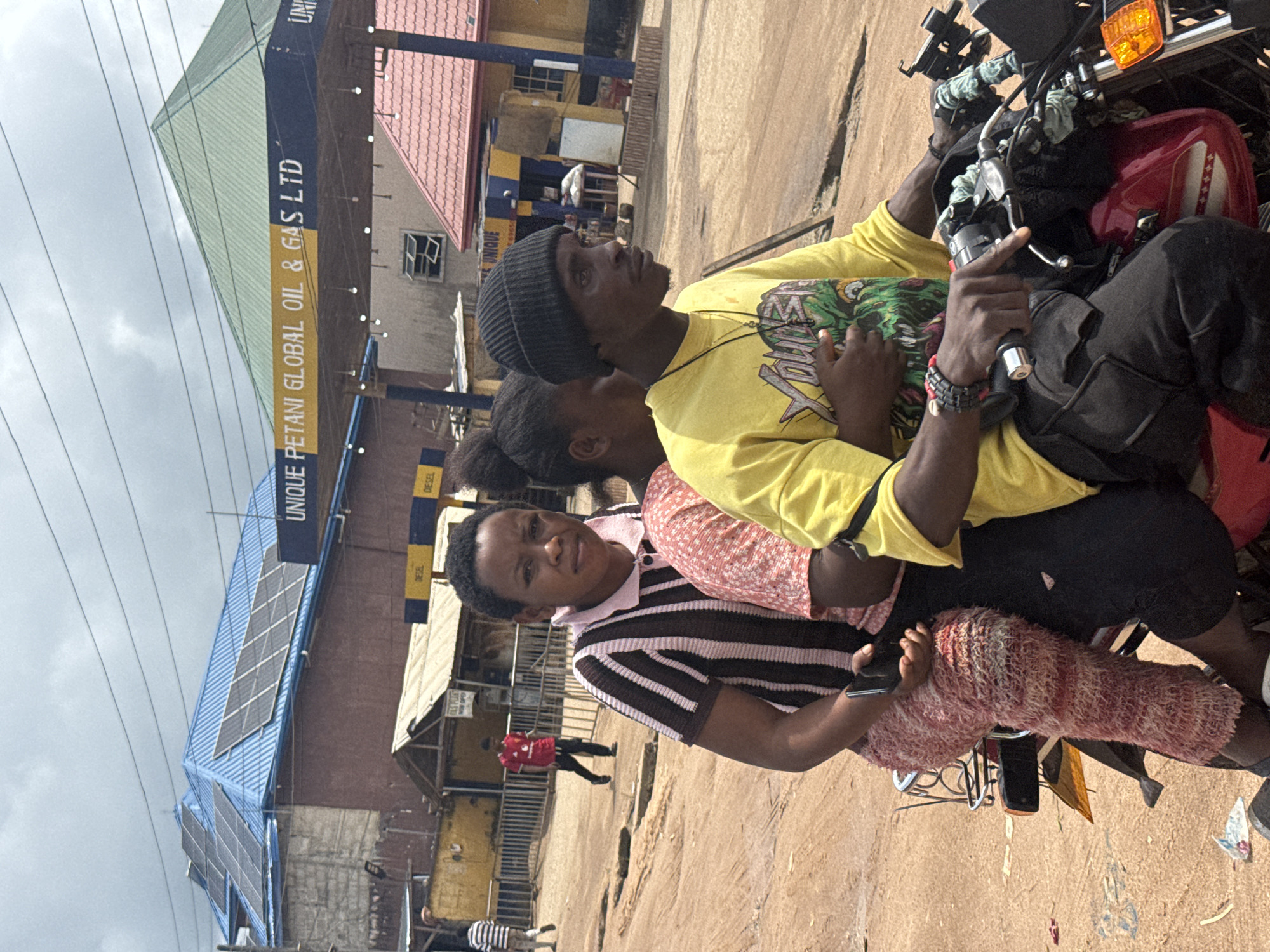 The image depicts a street scene in Ihiagwa, Nigeria. Three people are on a motorcycle: a man driving, wearing a yellow t-shirt and a black beanie; a woman behind him, wearing a pink and brown striped shirt and textured pink pants; and another person in the middle wearing a pink and white patterned top. The motorcycle is red and appears to be in motion on a dirt road. In the background, a gas station with a blue and yellow awning reads "UNIQUE PETANI GLOBAL OIL & GAS LTD". Buildings with corrugated metal roofs, some with solar panels, are visible. The sky is overcast. The road surface is uneven with visible divots.
