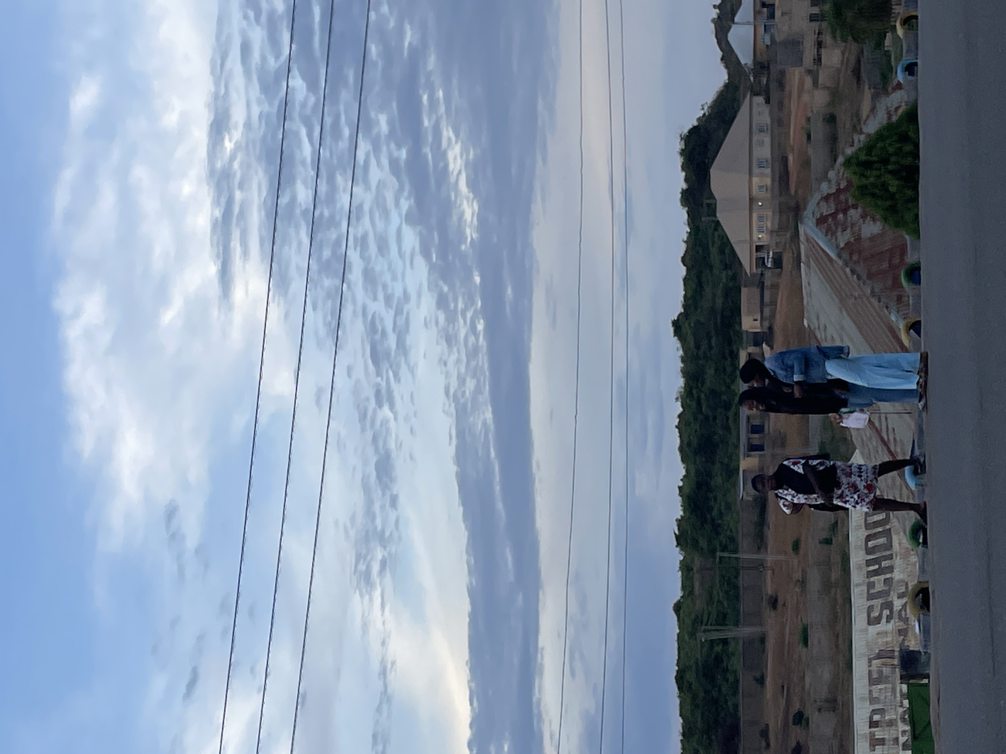 The image depicts a scene in Jos, Nigeria, with a focus on a group of people standing on a ledge overlooking a landscape. The sky above is partly cloudy, with several power lines cutting across the view. In the background, a building complex is visible, surrounded by trees and vegetation. The ledge itself is concrete, with "TREFOL SCHOOL" painted on it. There are three individuals visible on the ledge, all facing away from the camera. The first one is wearing a floral shirt and dark trousers. The next one is wearing a black shirt. The third person is wearing a denim jacket and a light-colored top and bottom.