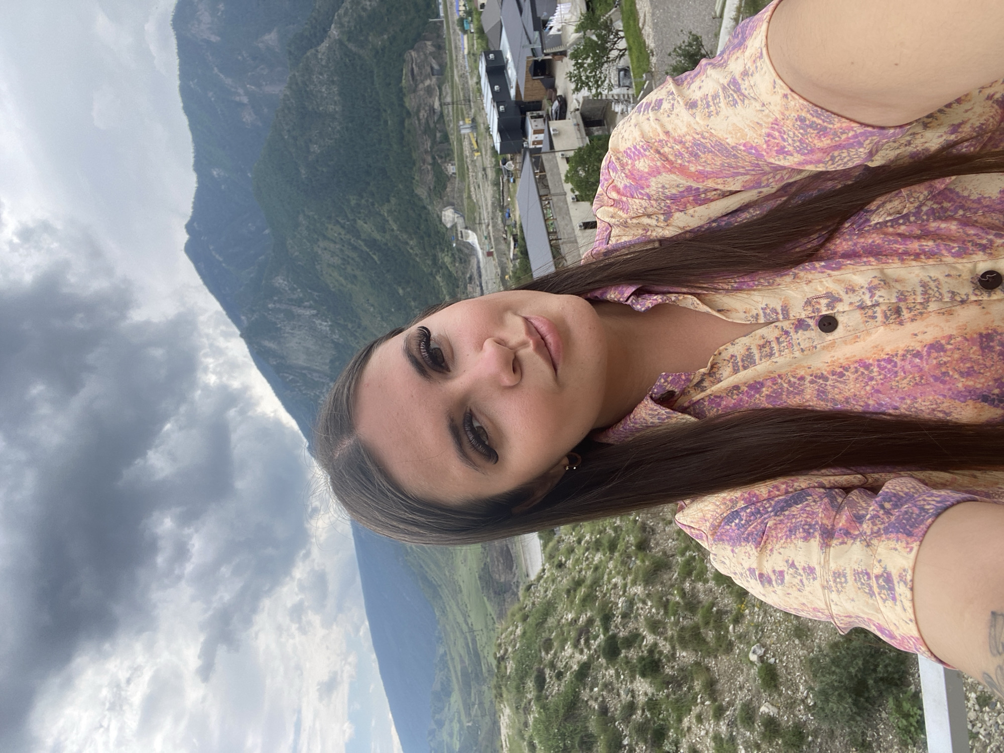 A young woman with long, dark hair is taking a selfie outdoors. She is wearing a light-colored shirt with a multi-colored abstract pattern. Her gaze is directed towards the camera, and she has a slight smile. In the background, a mountainous landscape unfolds under a cloudy sky. The scene appears to be in the vicinity of Chalda, Russia, with a small settlement visible in the mid-ground, nestled amongst the hills. The settlement features modern buildings with dark roofs, and some construction or infrastructure projects are apparent. The natural environment is characterized by rugged terrain with green vegetation covering the slopes.