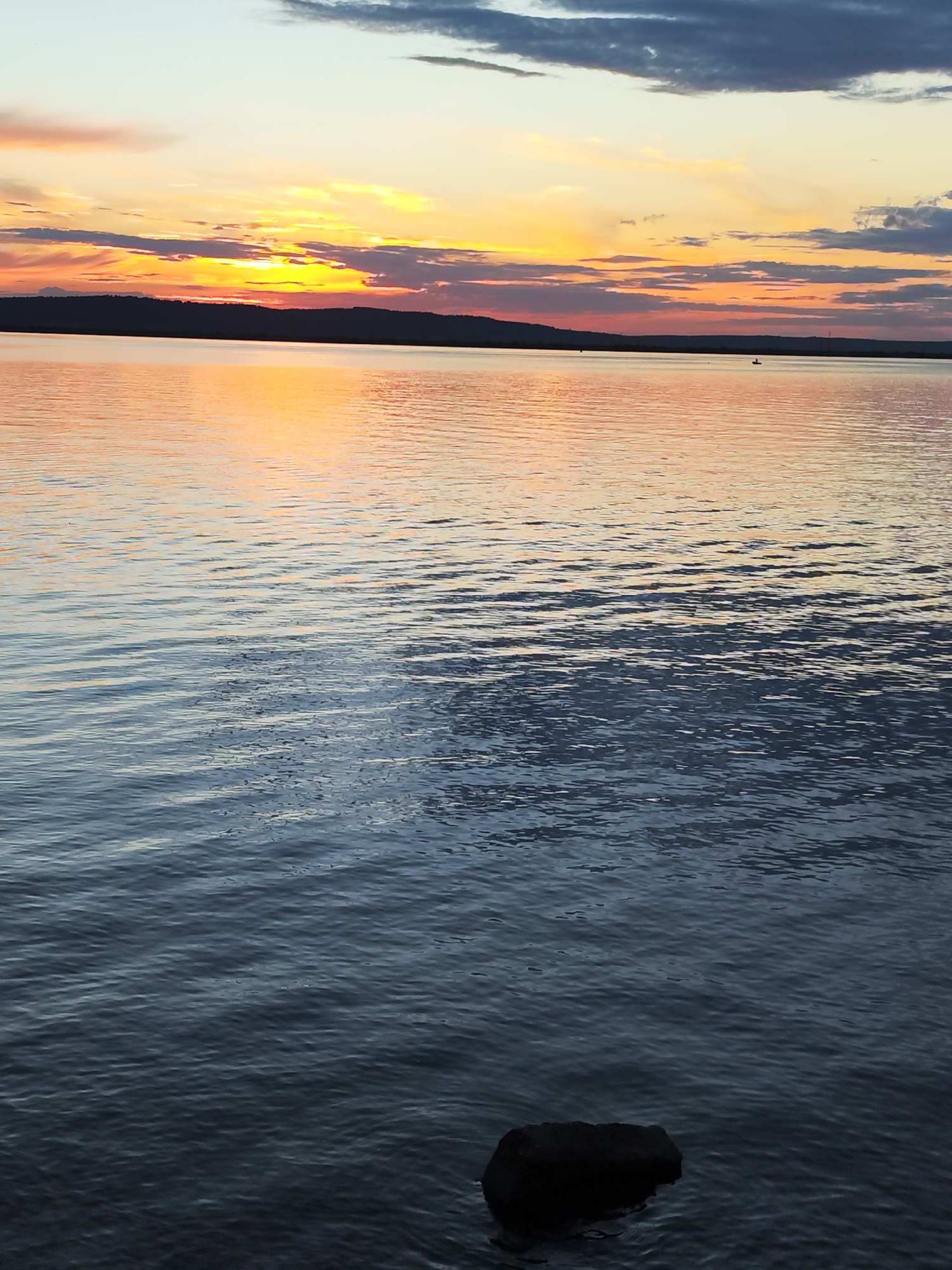 A picturesque sunset over the Kama River near Chaikovsky, Russia, is depicted. The sky is a vibrant canvas of orange, yellow, and pink hues, painted with streaks of gray and dark blue clouds. The sun's warm glow reflects on the calm water, creating a shimmering orange expanse that transitions to deeper blues and grays in the foreground. A dark, silhouetted shoreline with a hilly landscape stretches across the horizon. In the distance, a small, dark figure, likely a person in a boat, is visible on the water. A single dark rock rests partially submerged in the shallow water near the bottom right of the frame.