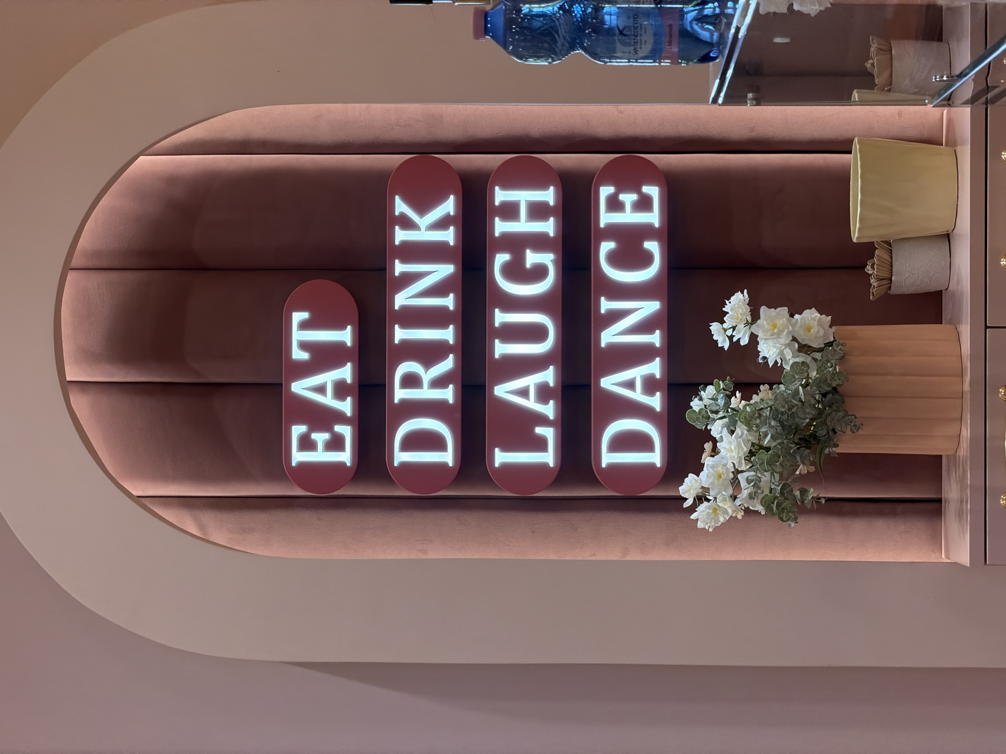 The image shows a brightly lit neon sign with the words "EAT," "DRINK," "LAUGH," and "DANCE" arranged vertically within red oval frames. The sign is set against a dark pink, tufted velvet background framed by a cream-colored archway. In the foreground, a vase of white flowers with green foliage sits on a light pink surface, possibly a shelf or counter, which has a mirrored reflection. To the right of the sign, a blue plastic bottle of San Benedetto water is visible. The overall aesthetic is modern and decorative, likely an interior space such as a cafe, restaurant, or a themed establishment. The location is identified as Sofia, Bulgaria.