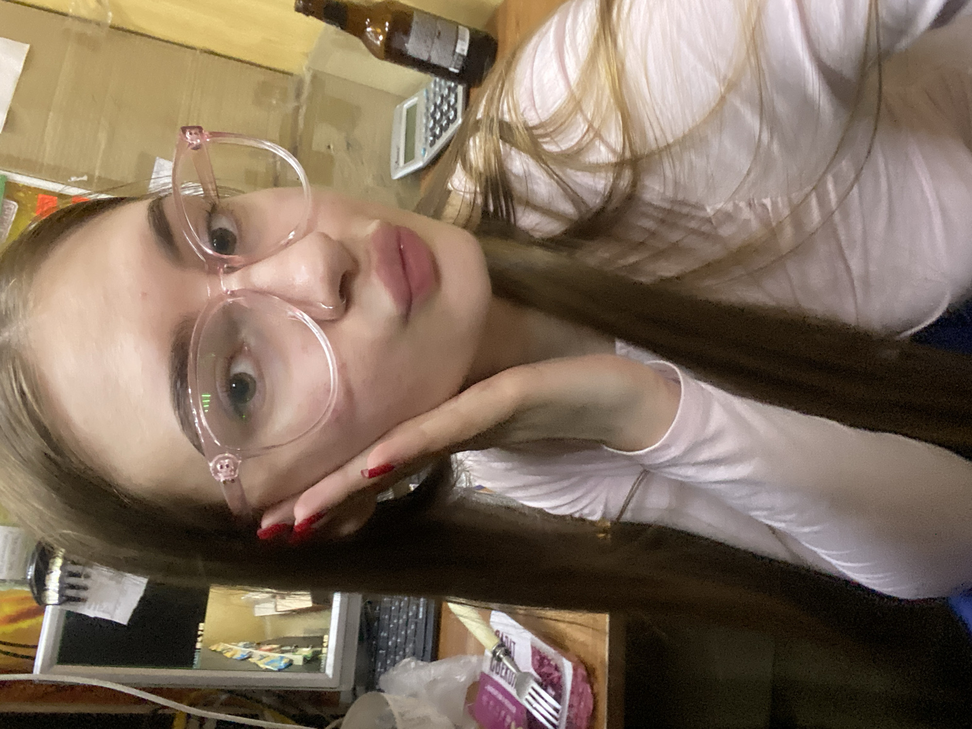 In a dimly lit room, a young woman with long brown hair and light-colored eyes is taking a selfie. She is wearing a light pink long-sleeved shirt and large, transparent pink-rimmed glasses. Her left hand is resting on her cheek, and her fingernails are painted red. Her lips are slightly pursed. On the wooden desk in front of her, there is a package of food with a fork on top, a calculator, and a brown glass bottle. In the background, a computer monitor and other office supplies are visible. The scene appears to be an indoor setting, possibly an office or a home desk area, in the city of Dyakovo, Russia.