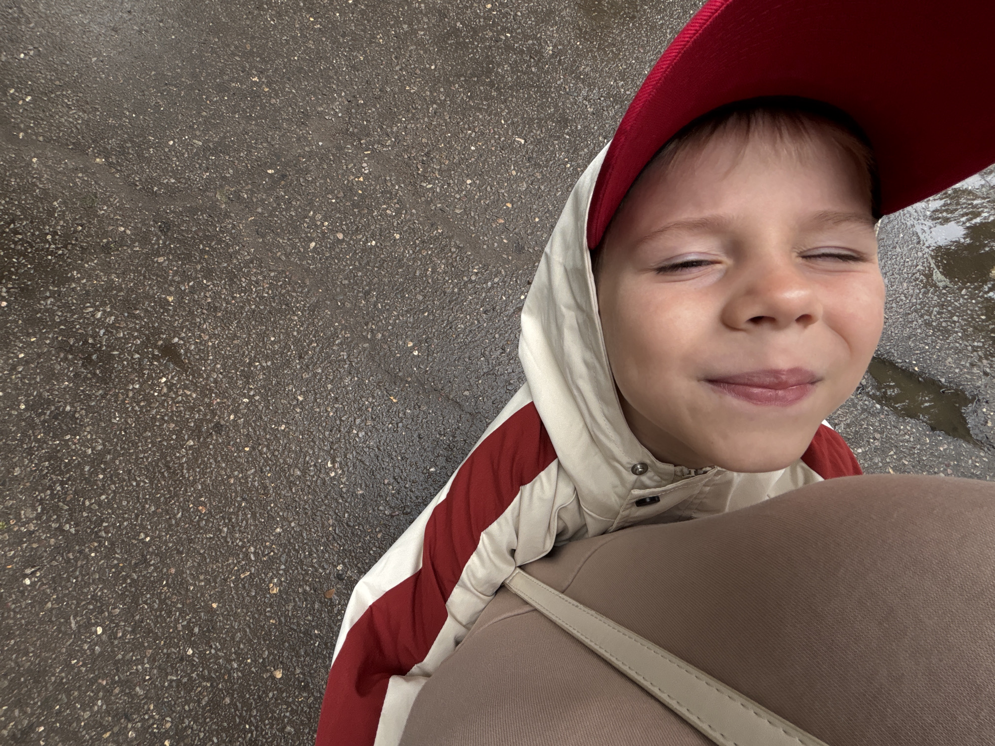 A child, likely under two years of age, is lying on a wet asphalt surface, their head tilted to the side and resting against a person's thigh or leg. The child is wearing a red hat and a jacket with red and white stripes. The child's eyes are closed, and their tongue is slightly protruding, suggesting a playful or tired expression. The person's leg is covered by light brown pants, and a beige strap from a bag is visible. The ground is dark gray asphalt, wet and speckled with small stones, indicating it may have recently rained. Puddles of water are visible on the ground. The scene appears to be outdoors, possibly in a park or a street in the city of Rusanovo, Russia. The overall lighting is muted, suggesting an overcast day.