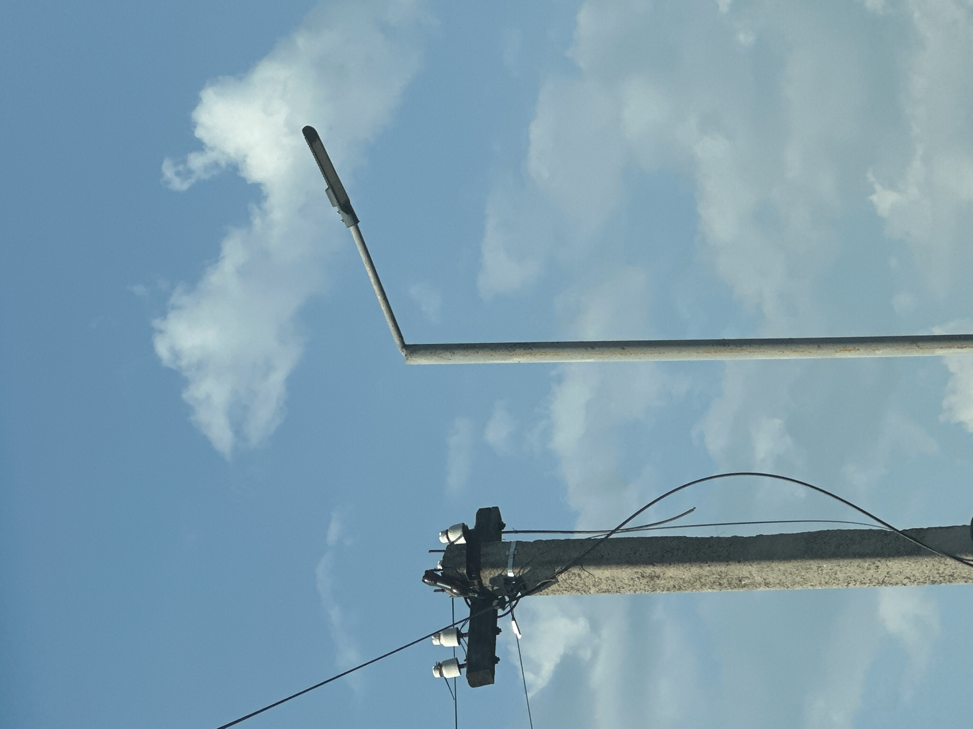 The image, taken in Shatava, Ukraine, shows a concrete utility pole with white insulators and electrical wires on the left, and a grey street lamp on a pole on the right, against a backdrop of a blue sky with scattered white clouds. The utility pole is made of rough concrete and has a horizontal wooden crossarm supporting three white ceramic insulators and electrical wires. There are also loose, dark wires hanging from the pole. The street lamp is a modern, elongated fixture mounted on a tall, slender grey pole. The scene appears to be outdoors, with no people or other discernible subjects present. The lighting suggests it is daytime.