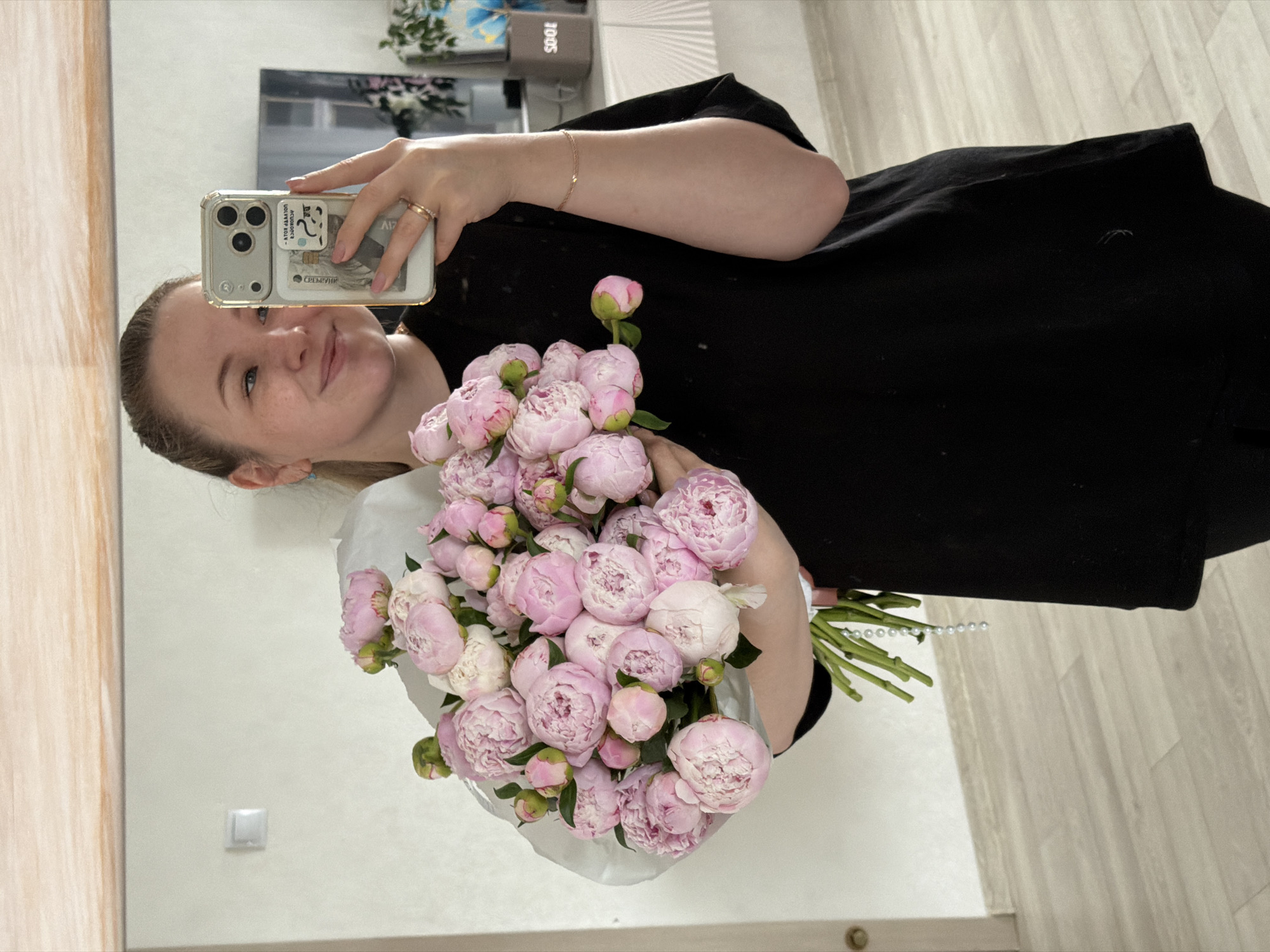 The scene is an indoor residential setting in the city of Chaikovsky, Russia. A young adult female, with blonde hair pulled back, is depicted in a mirror selfie. She is smiling, revealing light blue eyes. She wears a black short-sleeved top and dark lower garments. Her left hand holds a gold-colored smartphone, likely an iPhone, in a clear case with a multi-lens camera array. A clear card holder is attached to the phone's back, containing a credit card with inverted Cyrillic text (partially visible as 'БАНК' when correctly oriented) and a small, stylized sticker. A gold-toned ring is on her left ring finger, and a thin gold-toned bracelet is on her right wrist. A small light blue stud earring is visible in her left ear.
She holds a large bouquet of approximately two dozen light pink and white peonies, wrapped in light grey paper. Green stems and a decorative string of white pearls are visible below the wrapping.
The reflected background shows a modern interior with light-colored walls and a light wood-look plank floor. A white square light switch is on the left wall. To the right, a white cabinet with a ribbed front supports a grey rectangular object displaying 'SOOE' (inverted, "300S") and a dark plant arrangement. Above the cabinet, a dark-framed object, possibly a television or picture, is visible. The mirror has a light wood-colored frame at its upper edge.