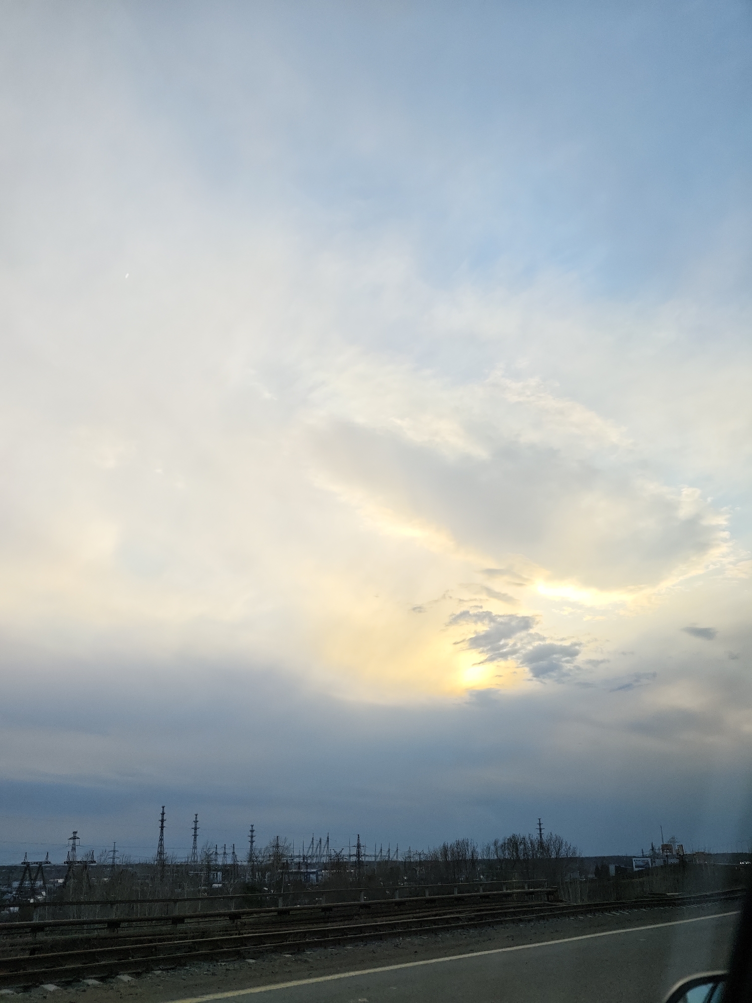 The image presents a wide view of an industrial landscape under an expansive, cloudy sky, likely captured from a moving vehicle. The sky occupies the majority of the frame, displaying a gradient from light blue at the top to various shades of white and grey, with a significant band of soft yellow and orange light breaking through dense clouds in the middle and right sections, indicating either late afternoon or early evening. A faint crescent moon is visible high in the sky in the upper-left quadrant.
Below the horizon, an extensive array of industrial infrastructure dominates the middle ground. Numerous tall power lines and transmission towers stretch across the scene, interspersed with bare trees and distant, indistinct industrial buildings. This suggests a cold season, such as late autumn or early spring.
In the foreground, multiple railway tracks run horizontally across the lower portion of the image. Parallel to the tracks on the right is a section of a paved road, implying the photograph was taken from a vehicle. A partial view of a vehicle's interior, including a side mirror and window frame, is visible in the bottom right corner. The scene evokes a sense of journey through an industrial area under a vast, dynamic sky, with no discernible text visible. The location context of Chaikovsky, Russia, aligns with the industrial and temperate landscape depicted.