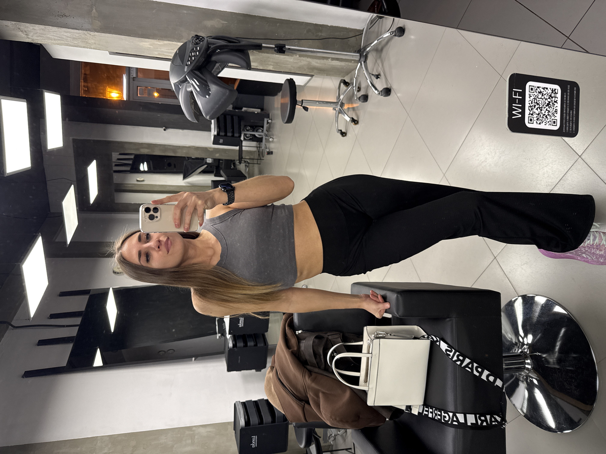 A woman with long brown hair is taking a selfie in a hair salon. She is wearing a gray cropped tank top and black leggings. She is holding a smartphone in her left hand. The background shows a hair salon with styling chairs, mirrors, and hair dryers. A black stool with a white purse and a brown jacket on it is near her. There is a QR code for WiFi displayed on a desk. It is not possible to determine the location in Saint Petersburg, Russia.