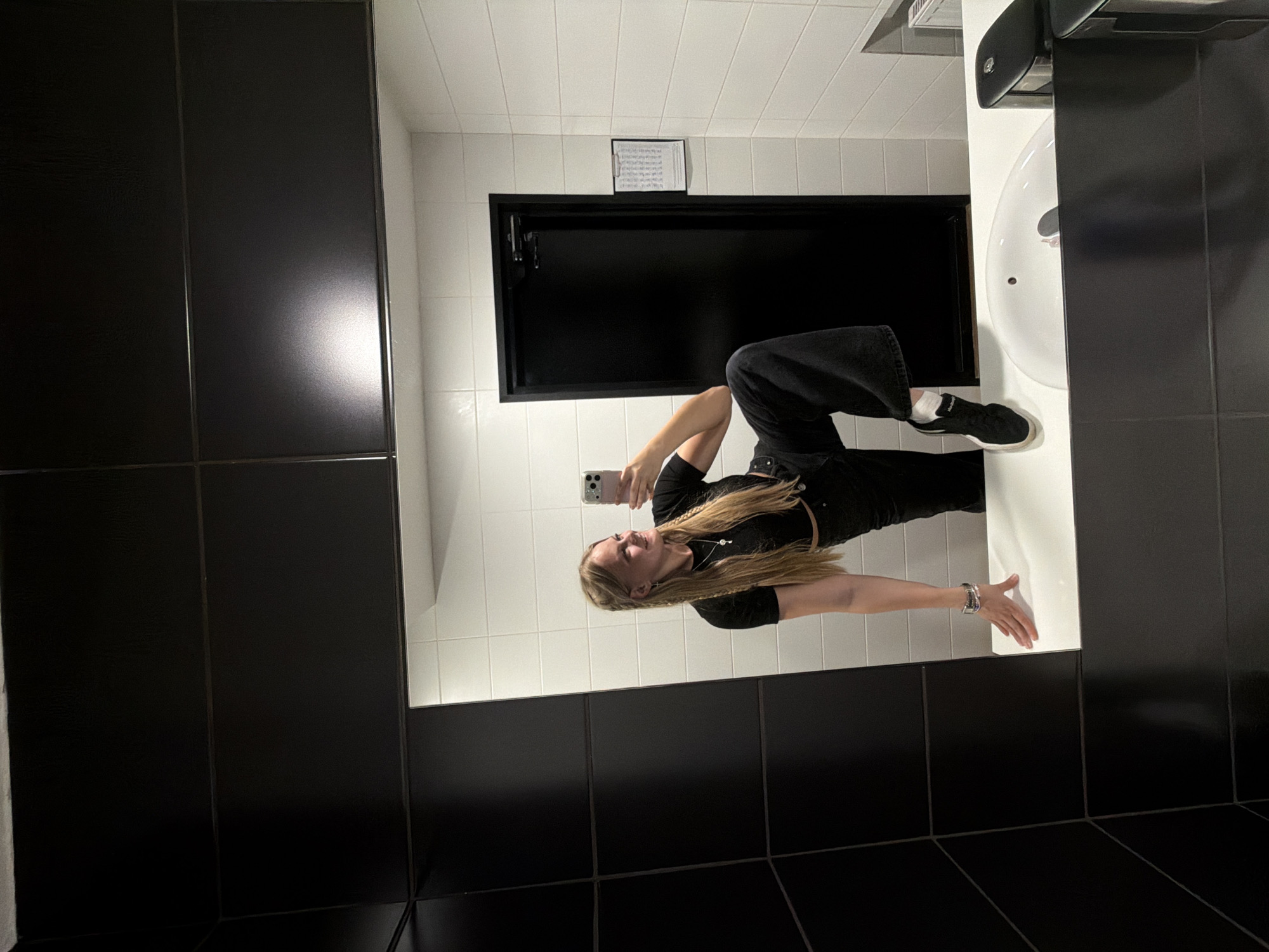 A young woman with blonde hair is taking a mirror selfie in a brightly lit, modern restroom. She is wearing a black crop top and black wide-leg pants, with white socks and black sneakers. Her hair is styled in braids, and she is wearing a silver necklace and bracelet. The restroom features white tiled walls and black tiled floors. A large black mirror reflects the scene. To the right of the mirror is a white sink with a chrome faucet. Above the sink, there is a black paper towel dispenser. Behind the woman, a black rectangular cabinet is visible on the wall, with a small framed document hanging above it. The overall impression is one of a stylish and clean public restroom. The lighting suggests it could be daytime or well-lit artificial lighting at night.